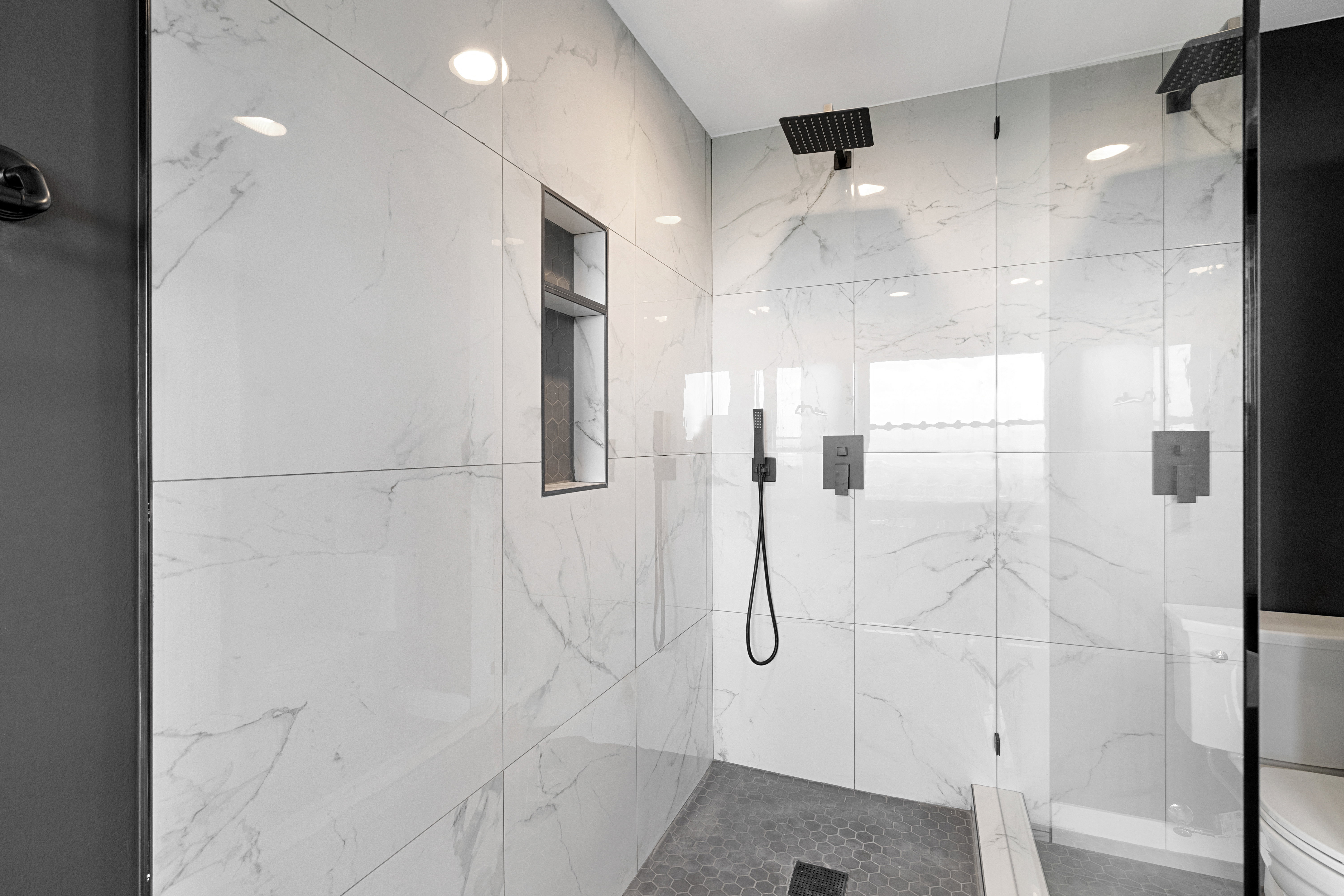 Spacious walk-in shower featuring large format white marble-look wall tiles, a built-in niche, and dual matte black rain showerheads