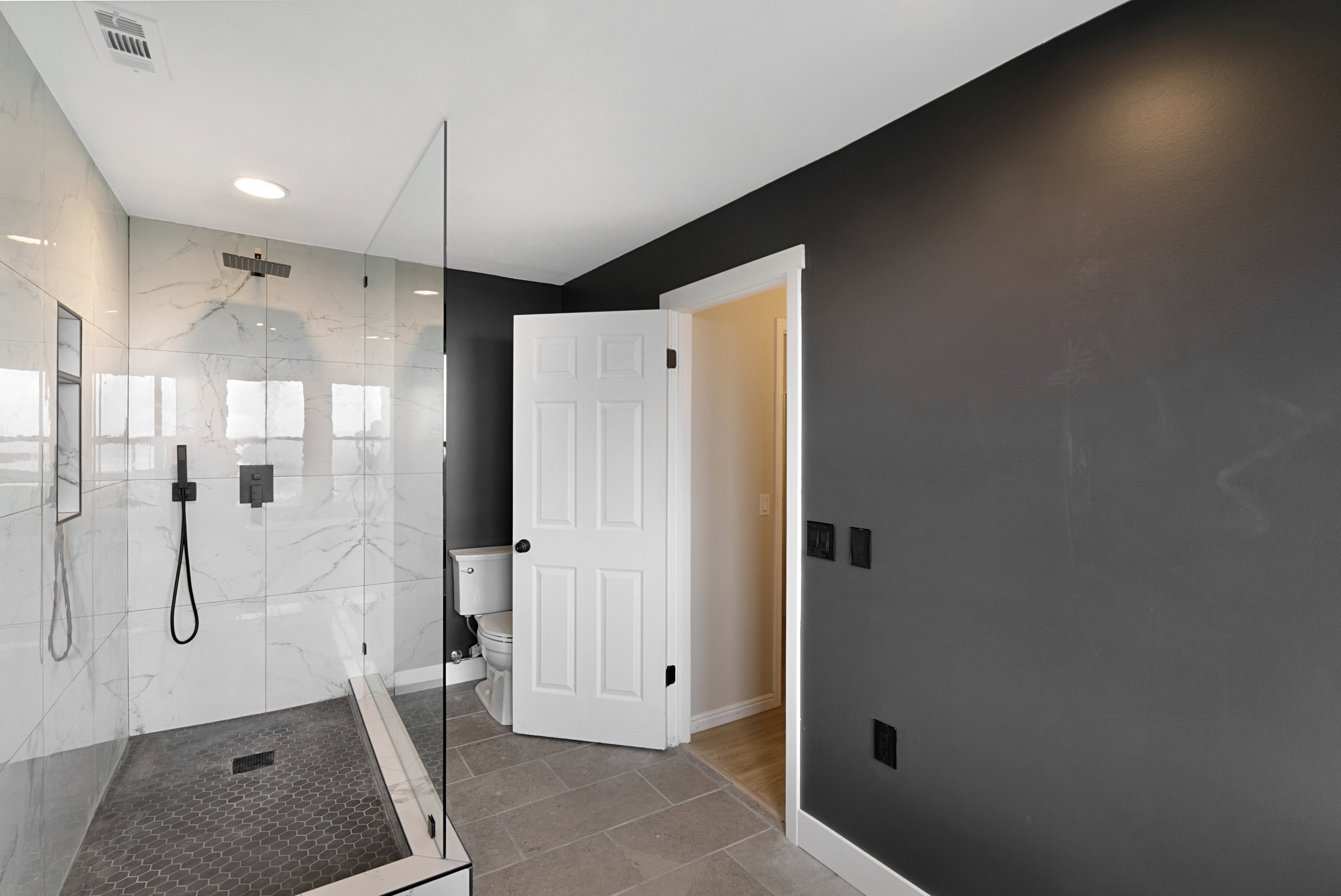 Spacious shower enclosure with clear glass paneling, white marble-style wall tiling, and dark hexagonal floor tiling
