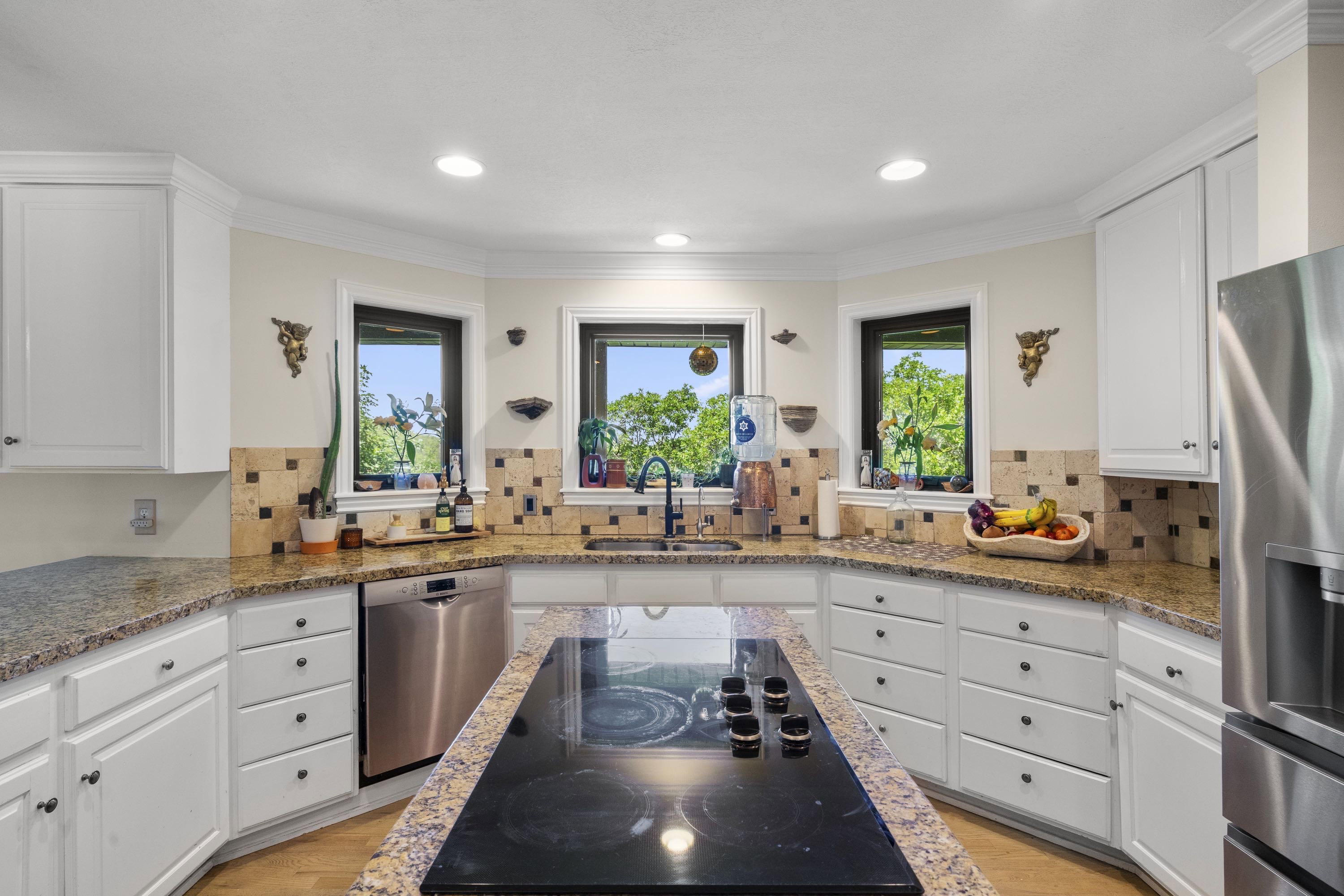 Spacious kitchen featuring an island with a built-in cooktop, white cabinetry, integrated stainless steel appliances, speckled countertops, and a tiled backsplash