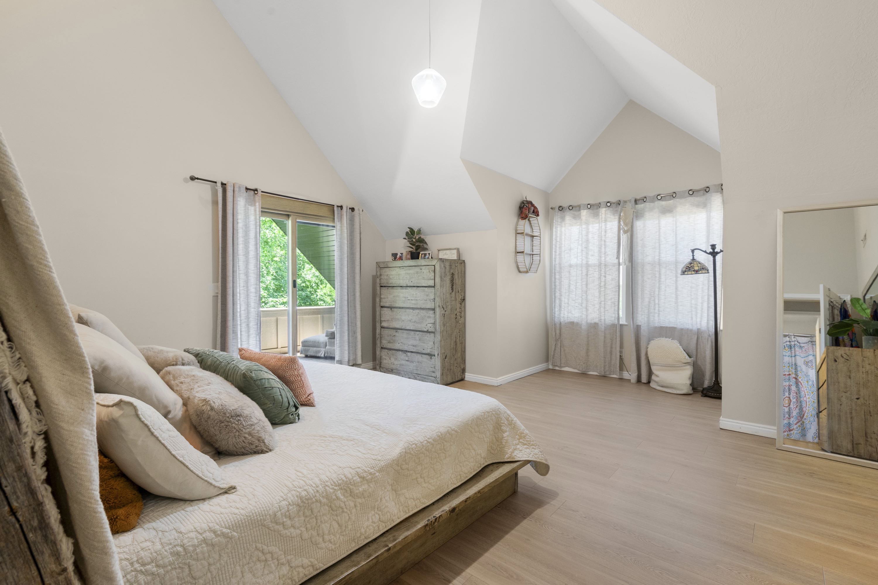Spacious room featuring vaulted ceilings, light wood-finish flooring, and a sliding glass door opening to an exterior balcony