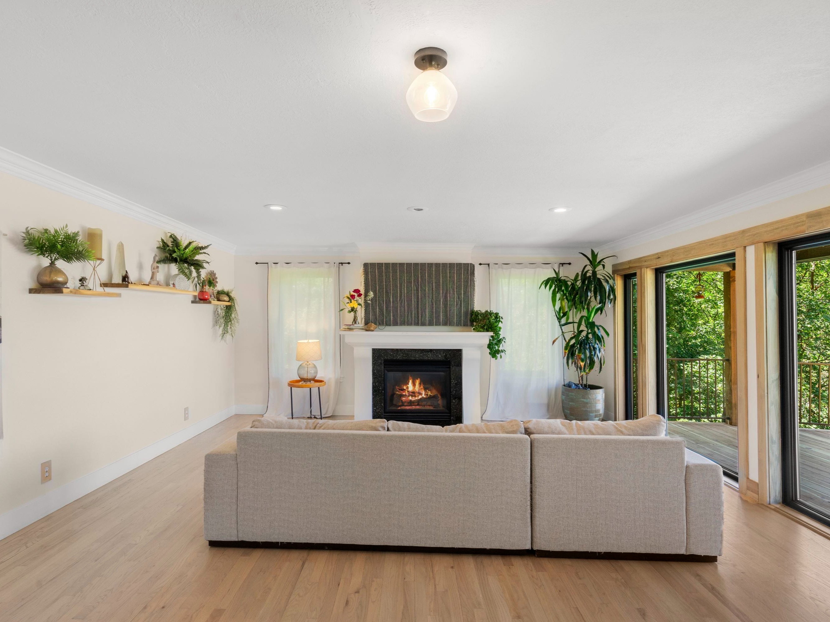 Spacious living area featuring light wood-finish flooring, a white mantel fireplace with a dark-tiled surround, crown molding, recessed lighting, and multiple sliding glass doors opening to a wood deck