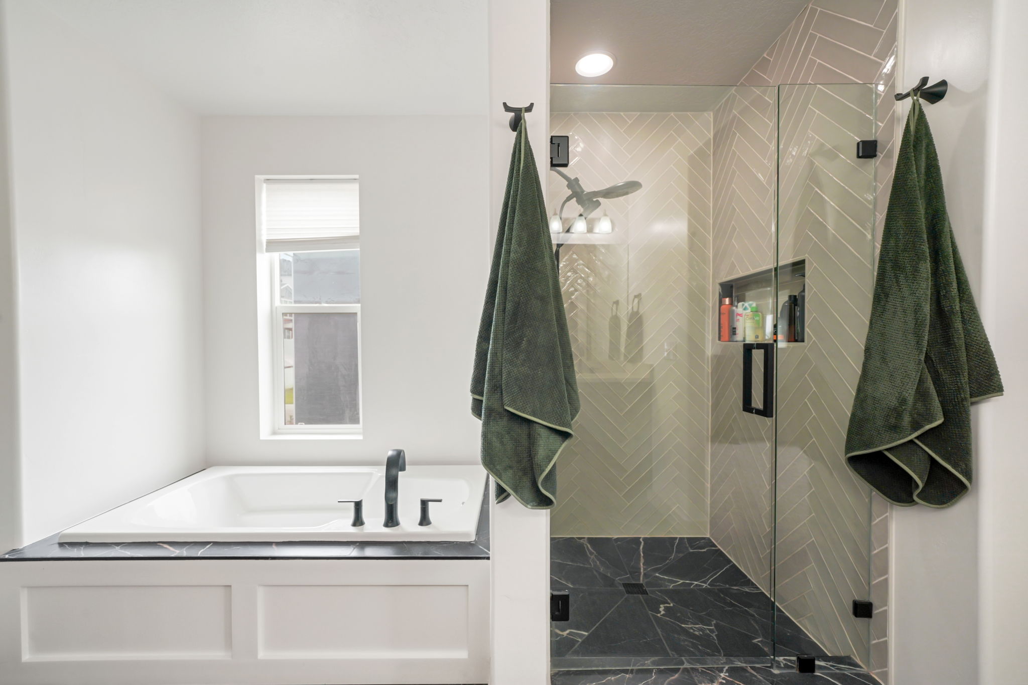 Modern bathroom featuring a built-in tub with a black countertop surround, a walk-in shower with clear glass doors, a chevron tile shower wall, and a dark stone-finish shower floor