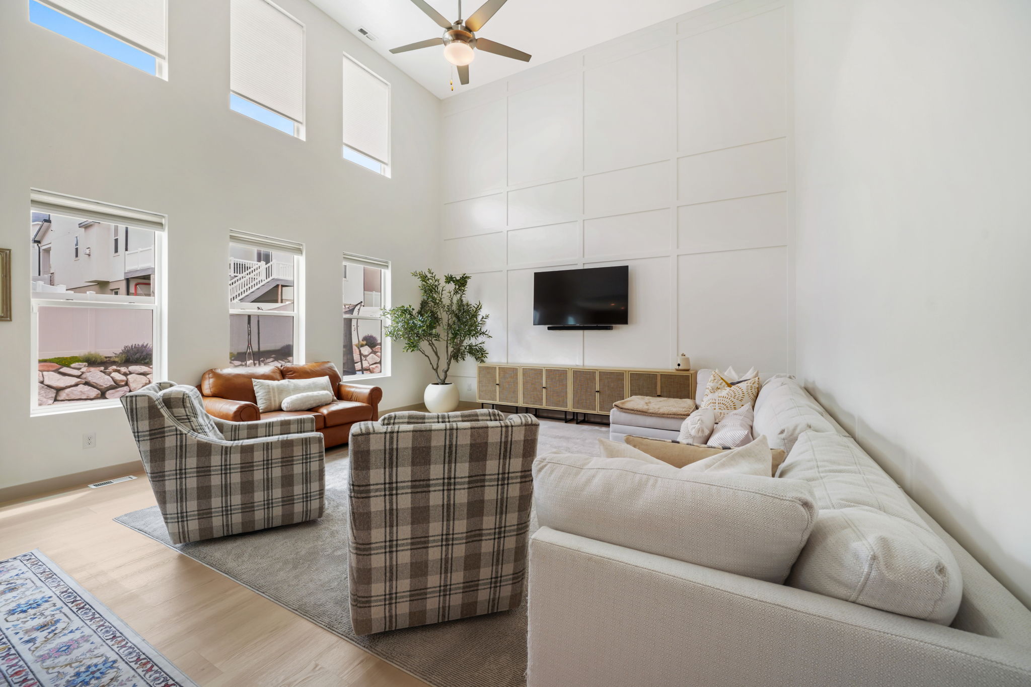 Vaulted living area featuring a decorative accent wall, ceiling fan, and high windows with roller shades