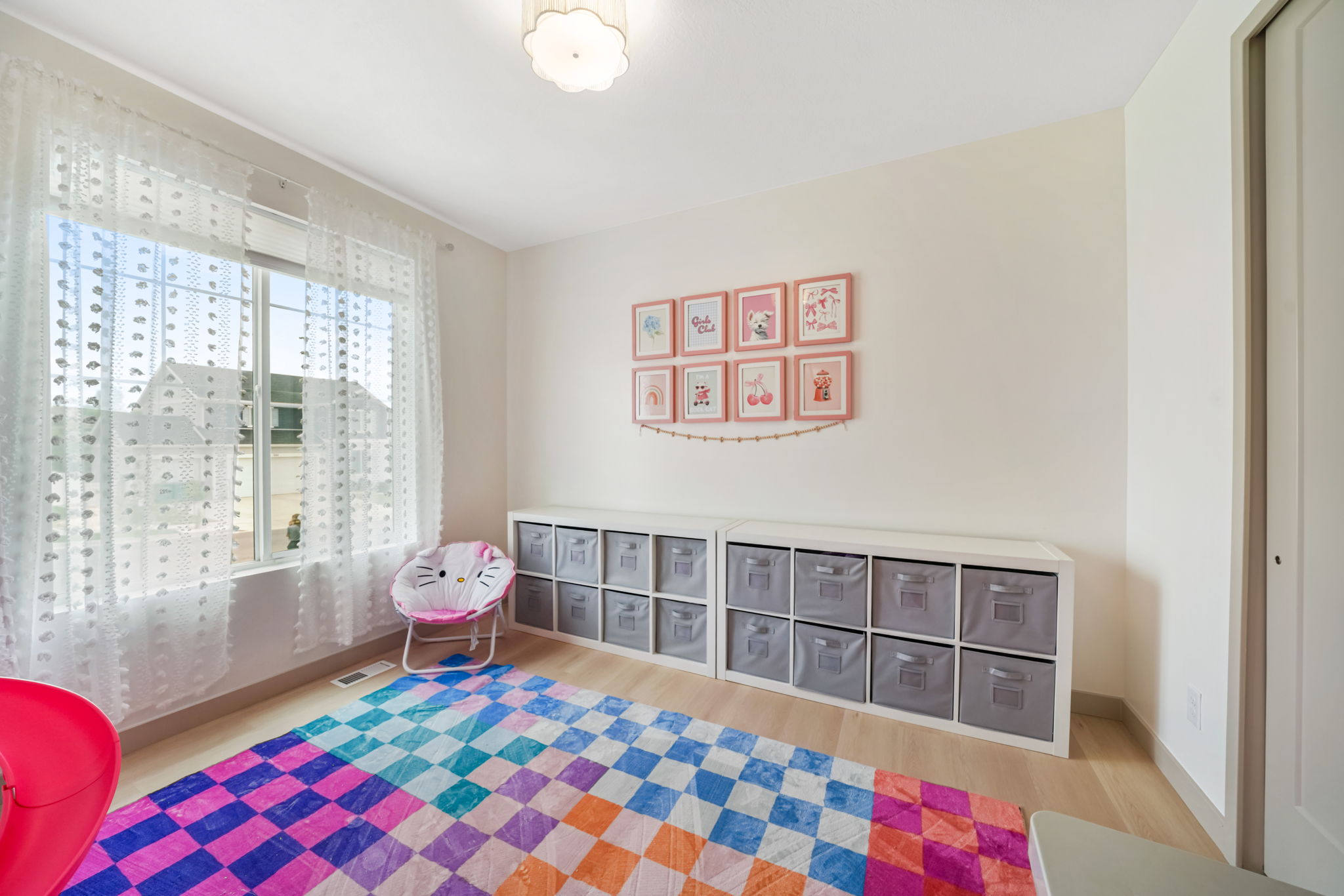 Bright room featuring a large window with sheer white curtains, light wood-finish flooring, a ceiling-mounted light fixture, and built-in storage cubbies