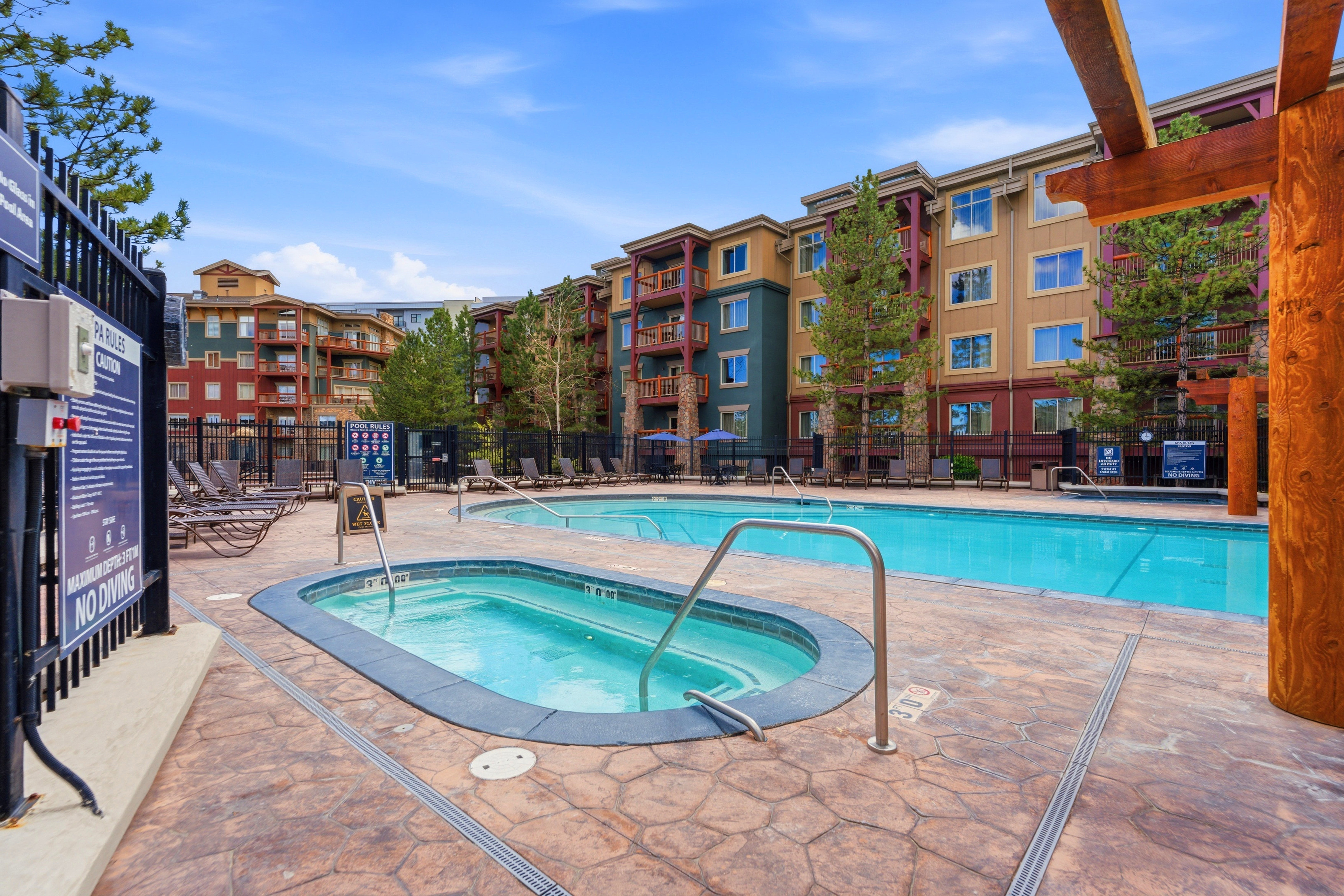 Outdoor pool and hot tub area featuring a stamped concrete deck, perimeter fencing, and adjacent multi-story buildings with balconies