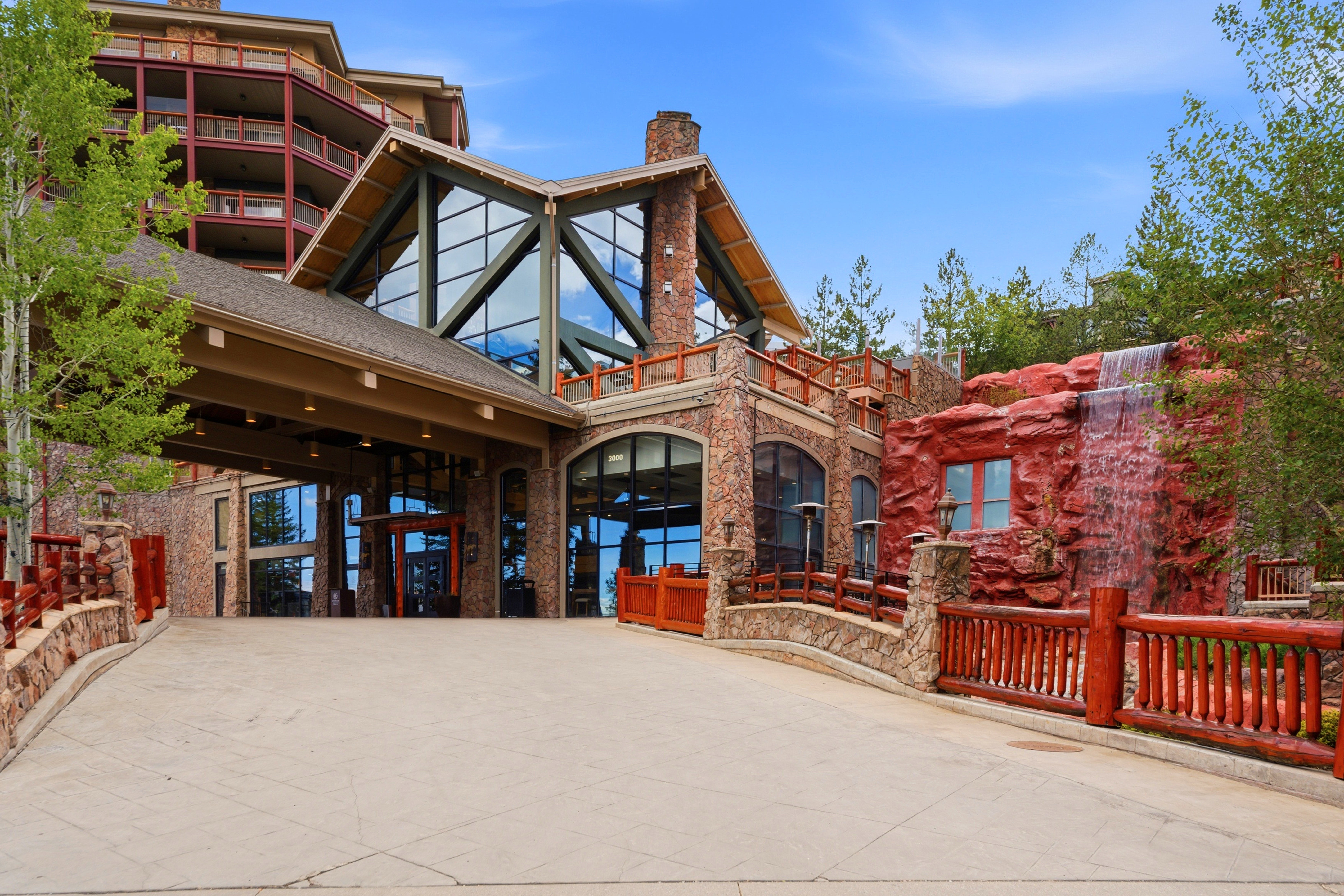 Grand entrance featuring a porte-cochère, stone facade, multi-story glass curtain wall, integrated water feature, and rustic log railings