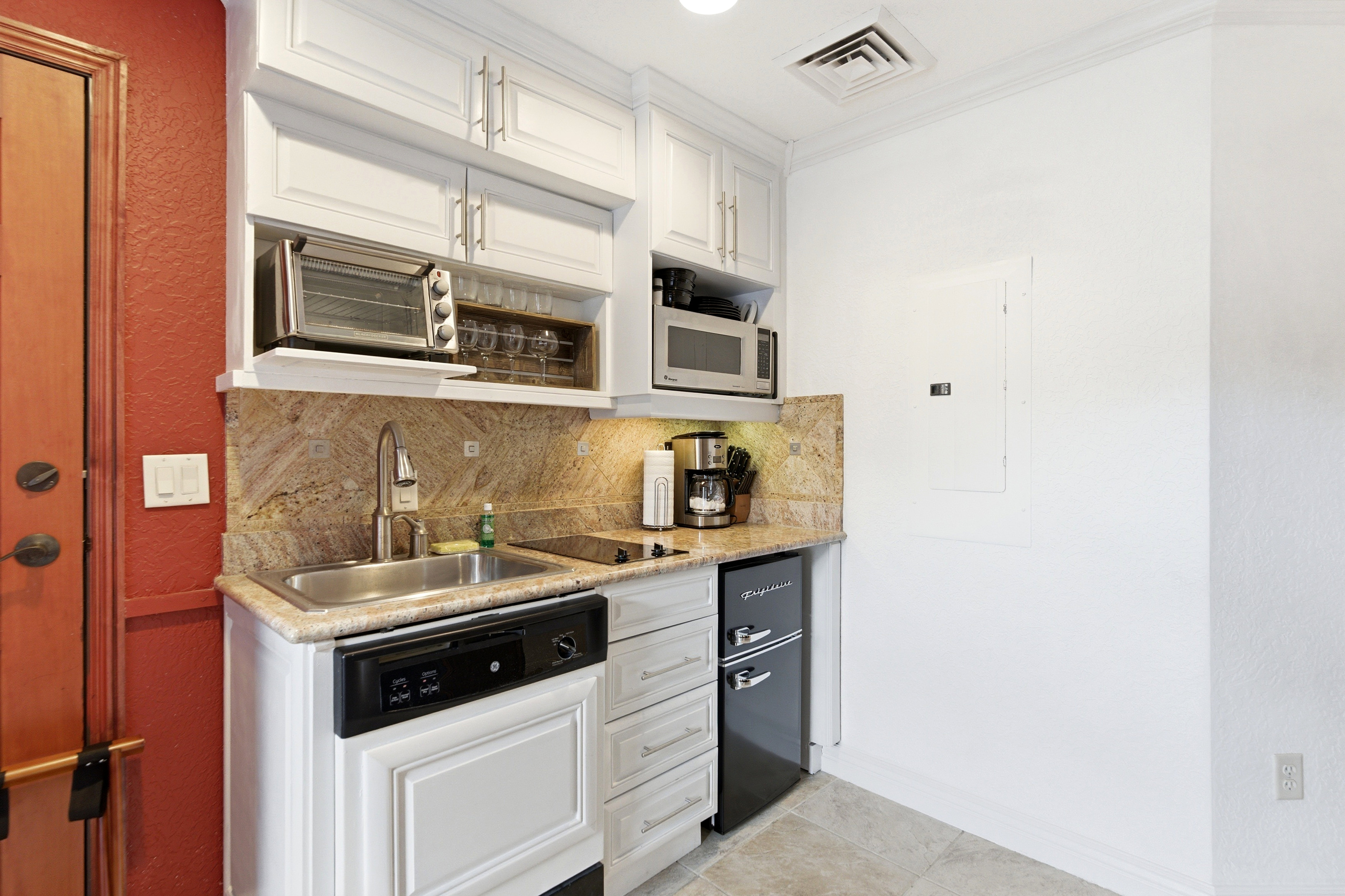 Compact kitchen area featuring white cabinetry, a stainless steel sink, a stone-finish backsplash, and an integrated dishwasher