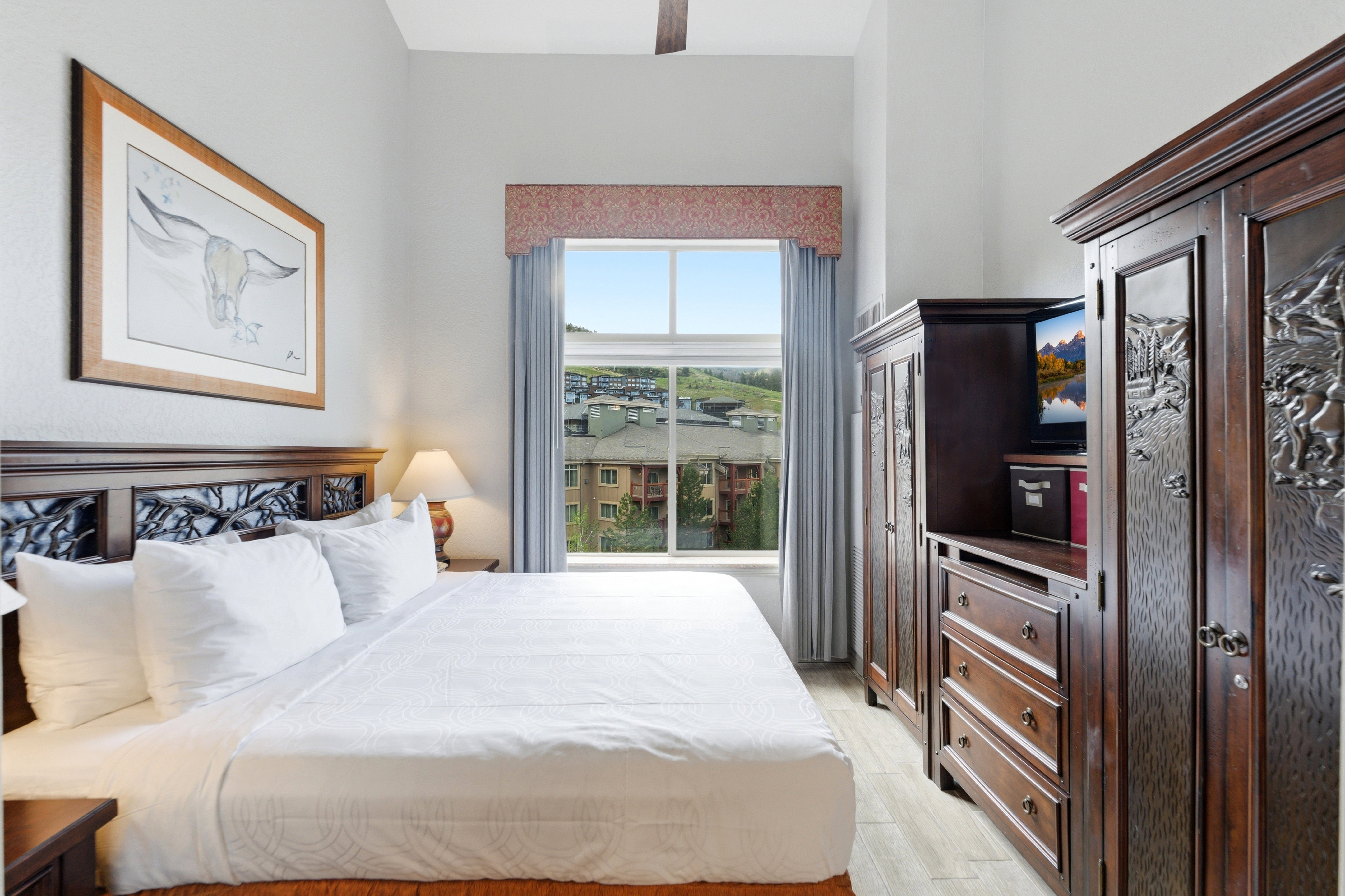 Room featuring wood-finish flooring, a window with drapery, a ceiling fan, a dark wood headboard with carved details, and a dark wood armoire system with carved panels and integrated storage