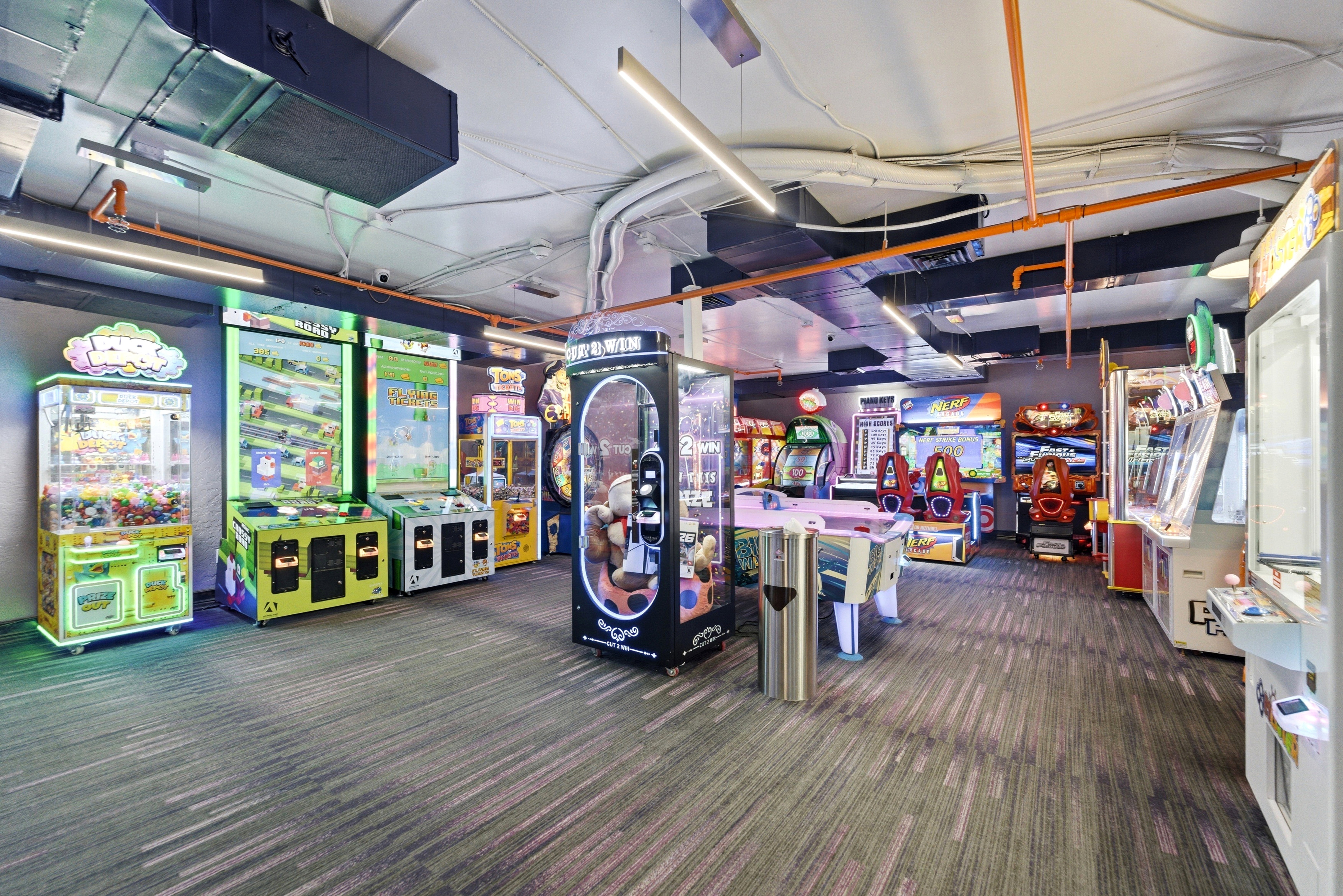 Spacious entertainment area featuring patterned carpet flooring, exposed ductwork, fluorescent lighting, and a variety of arcade-style game machines