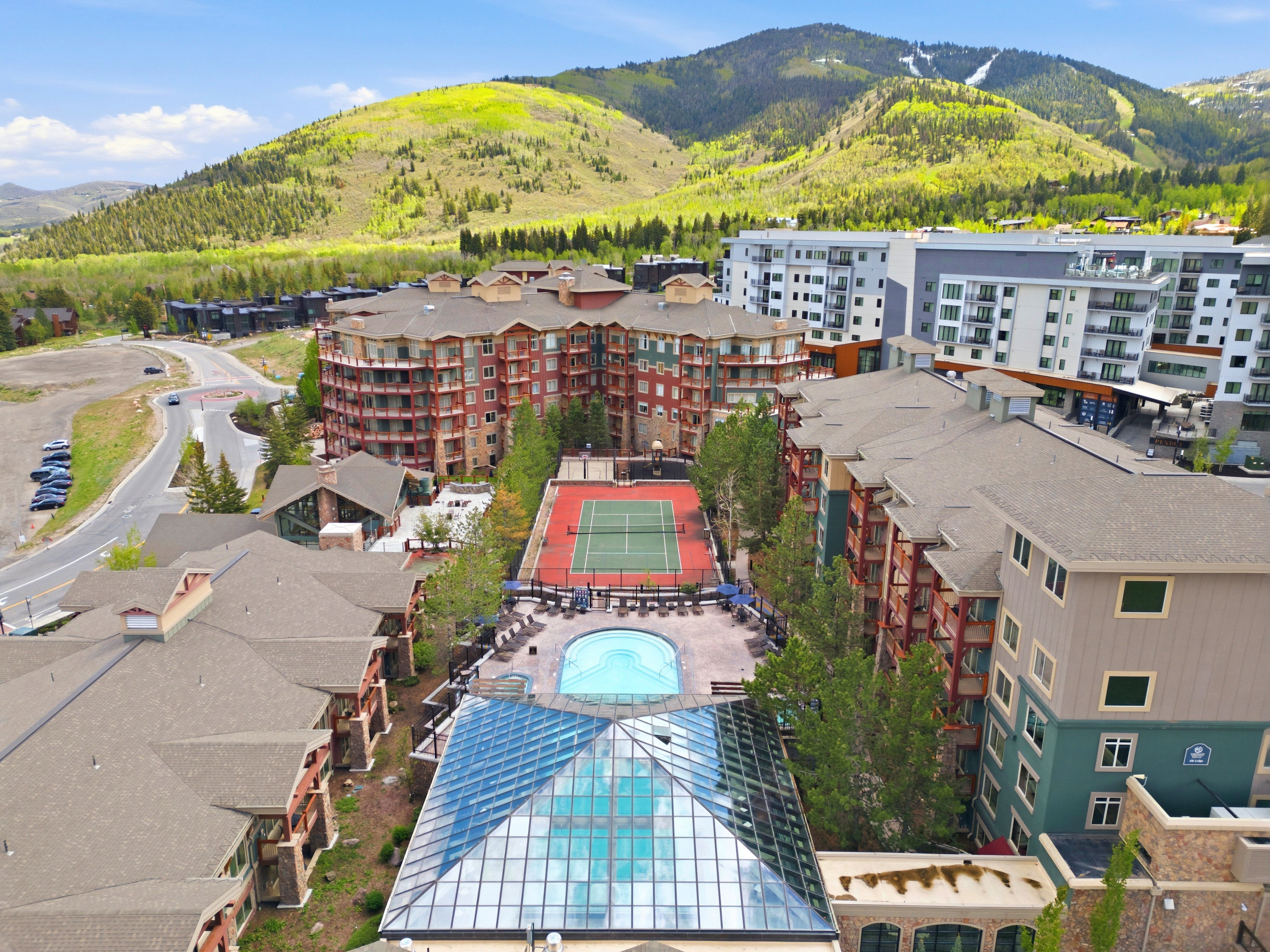 Aerial view of a multi-building complex featuring a central outdoor swimming pool, tennis court, and an indoor pool with a glass pyramid enclosure