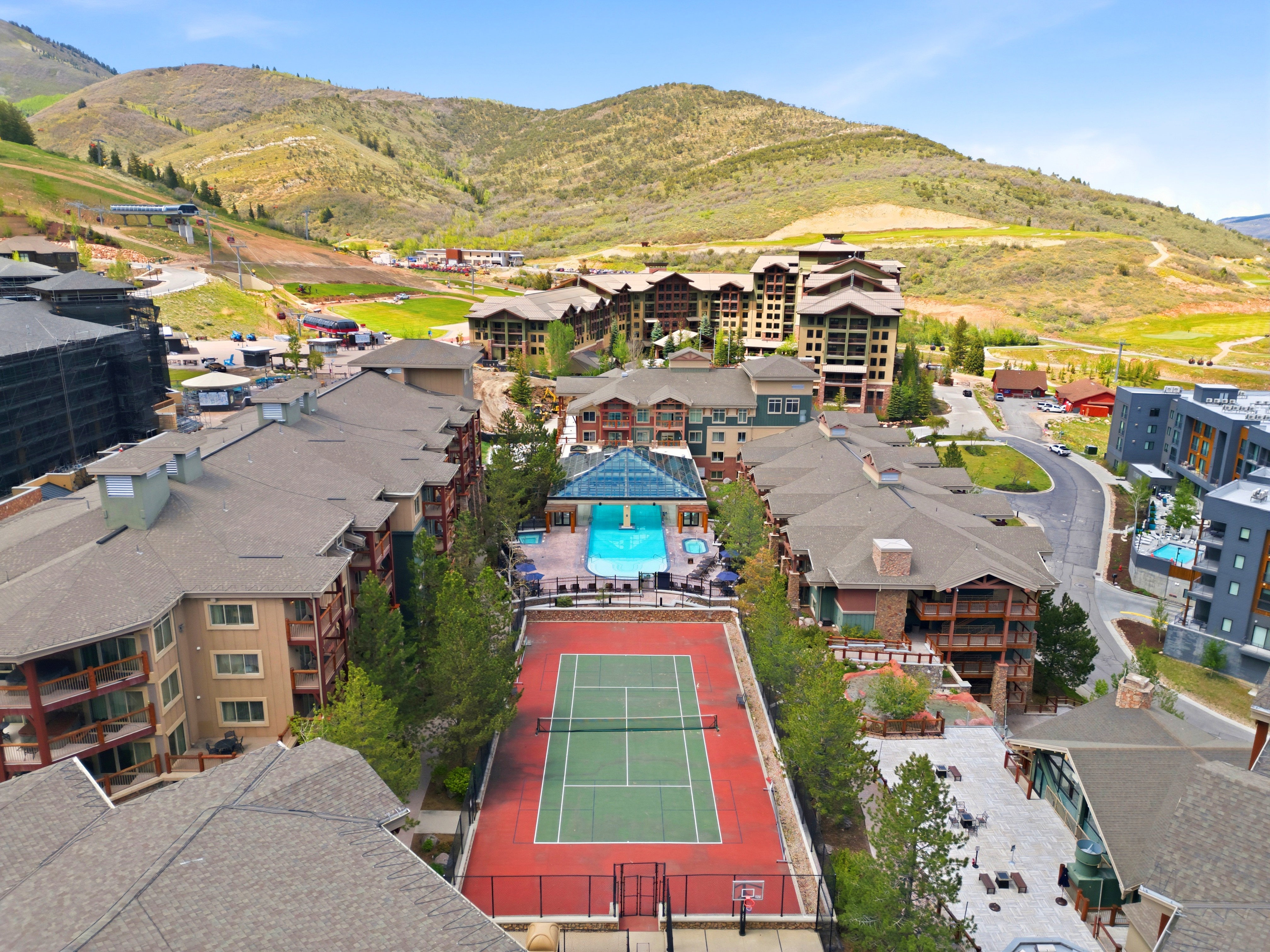 Aerial perspective showcasing a tennis court, an outdoor swimming pool, surrounding buildings with balconies, and distant mountains