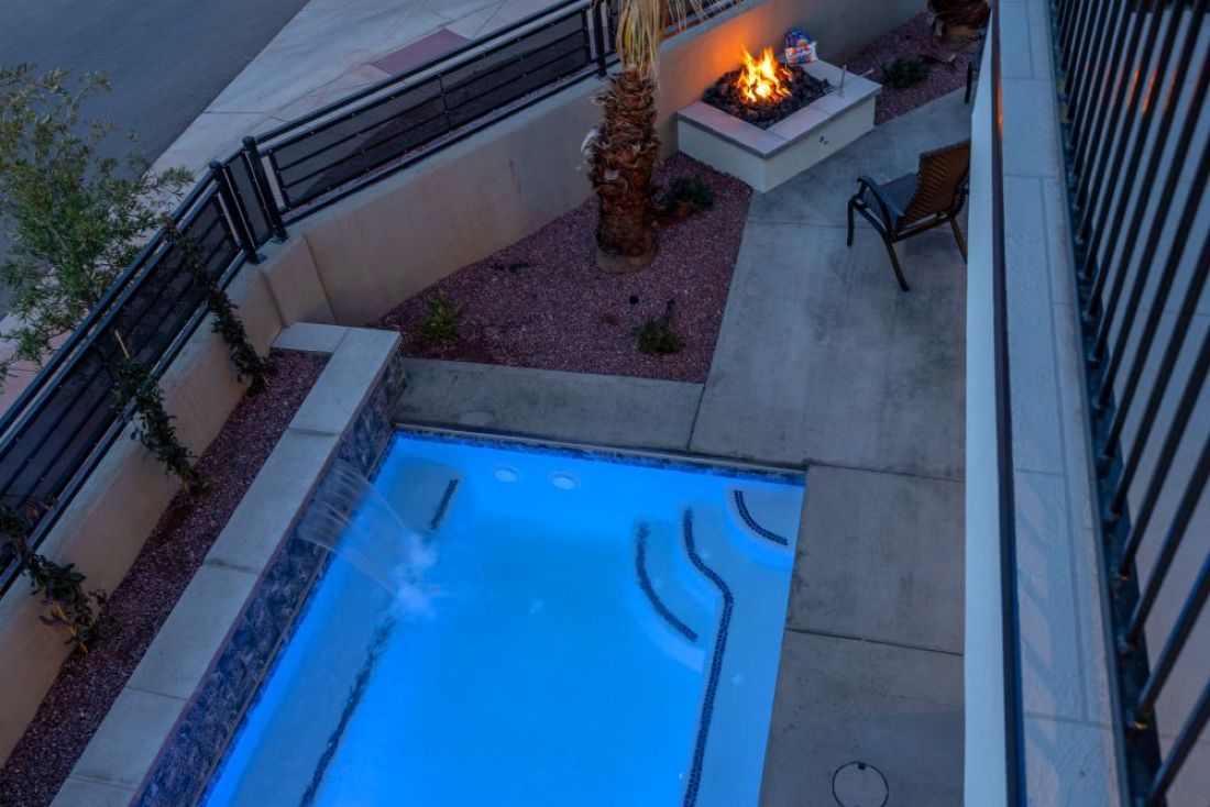 Outdoor space featuring a luminous swimming pool with a waterfall feature, a built-in fire pit, paved patio areas, and a rock-landscaped bed with a mature palm