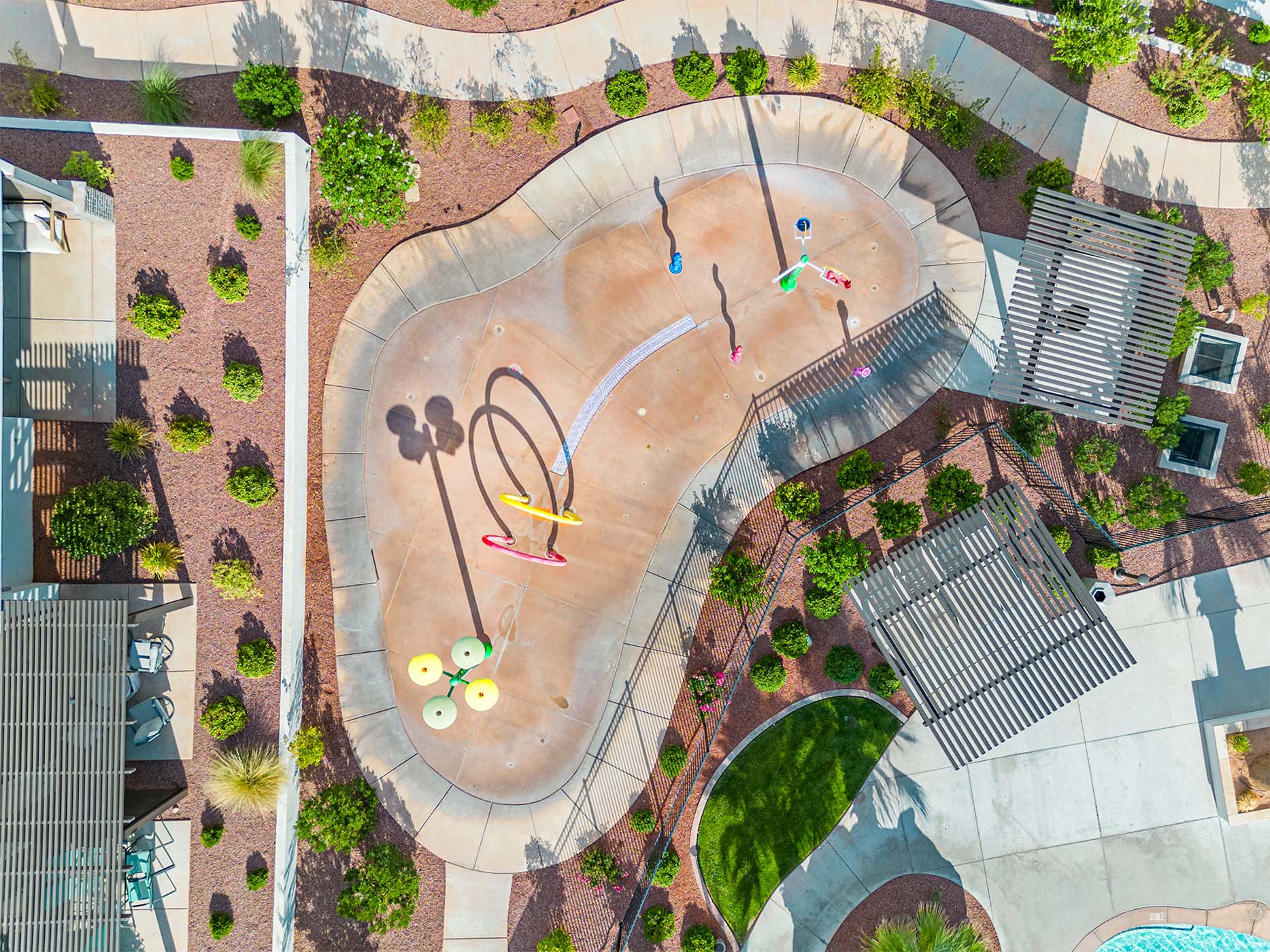 Splash pad with multiple water features, surrounded by pathways, two slatted pergolas, and landscaped grounds with mature shrubbery and decorative rock