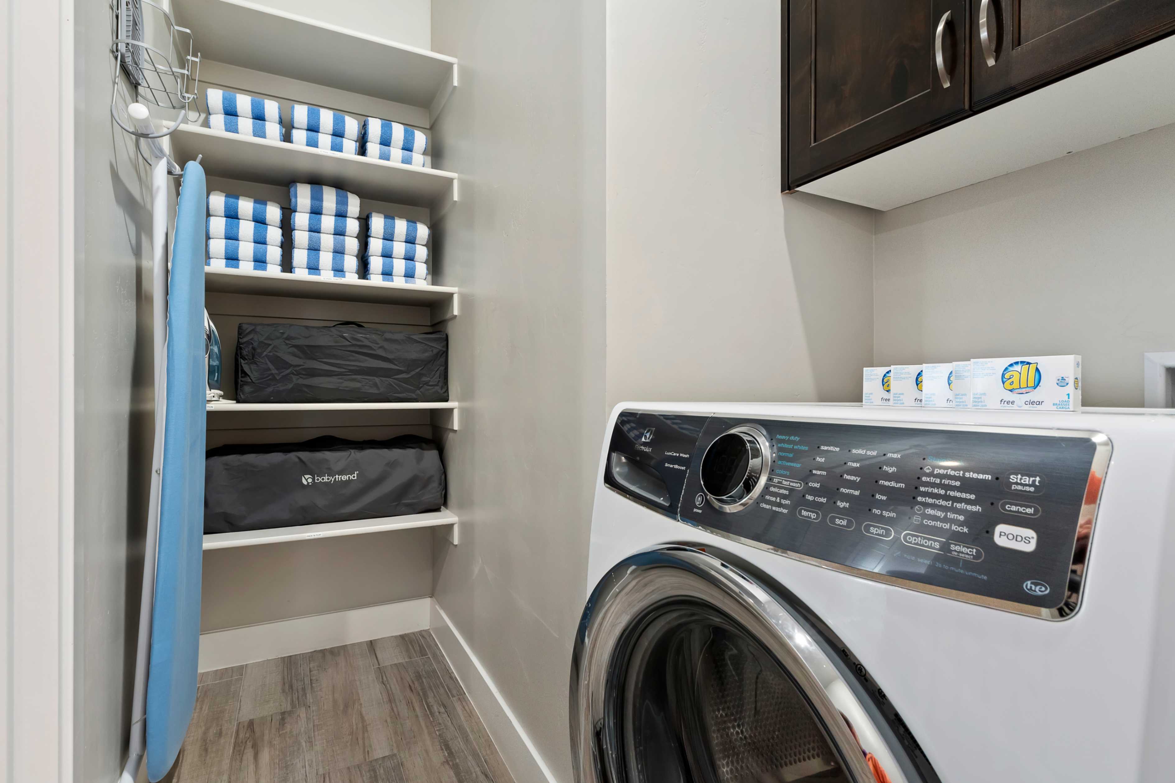 Dedicated laundry area featuring a front-loading washing machine, built-in shelving, dark wood-finish upper cabinetry, light gray wall paint, and wood-finish flooring