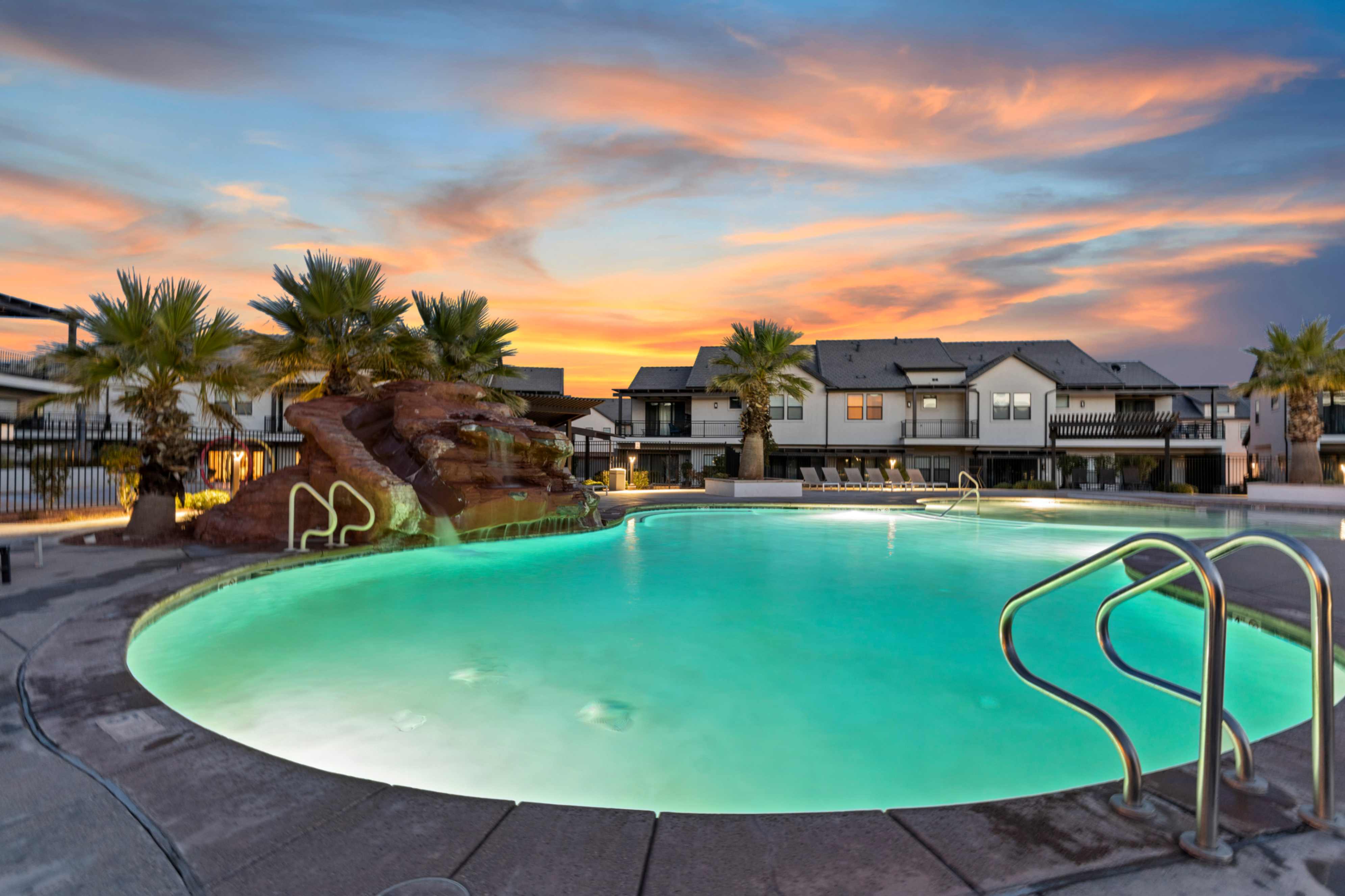 Curved swimming pool featuring a rock waterfall, mature palm trees, and perimeter concrete decking