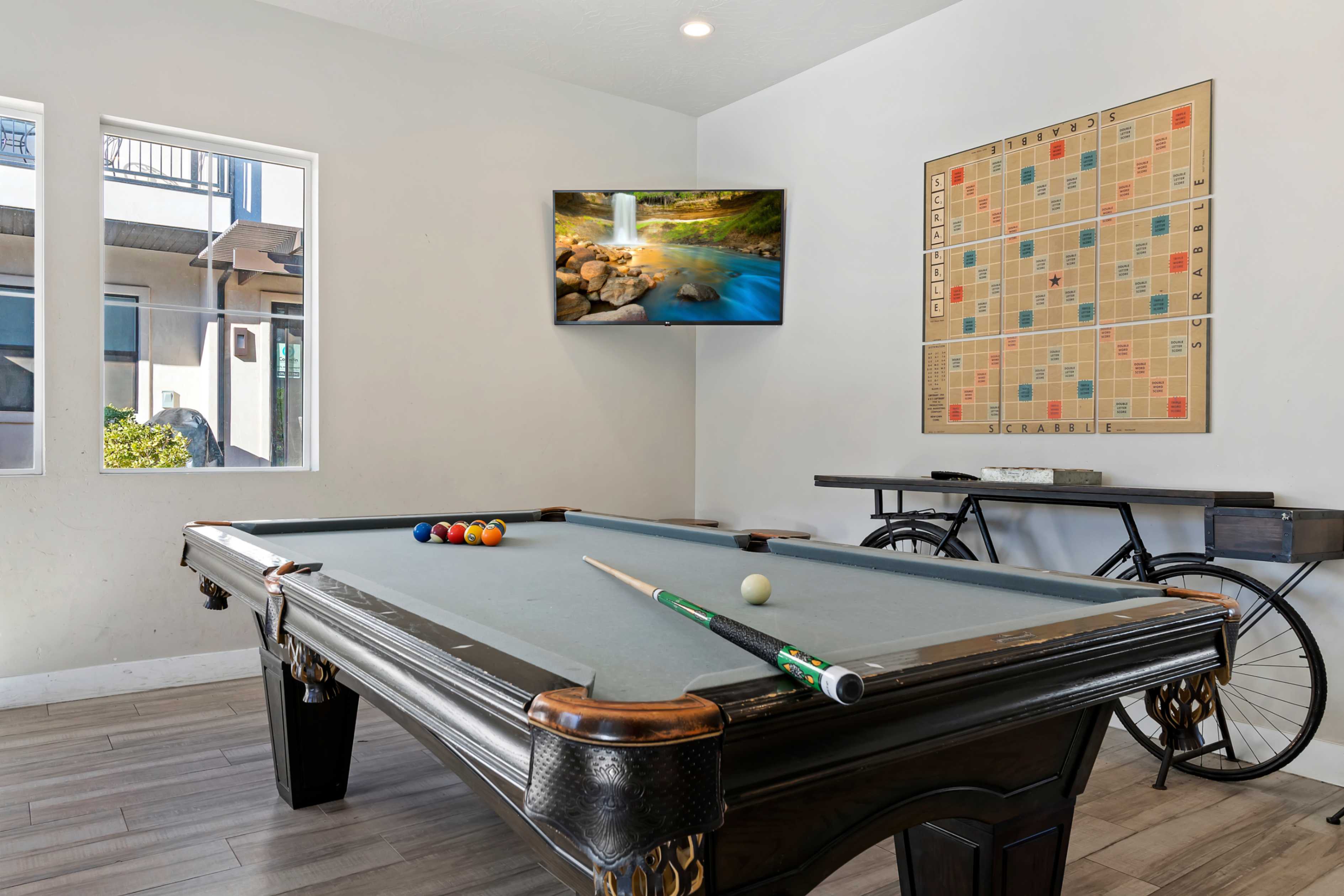 Recreation room featuring a classic billiards table with a light grey felt surface, wood-finish flooring, a wall-mounted flat-screen television, and multiple large windows for natural illumination