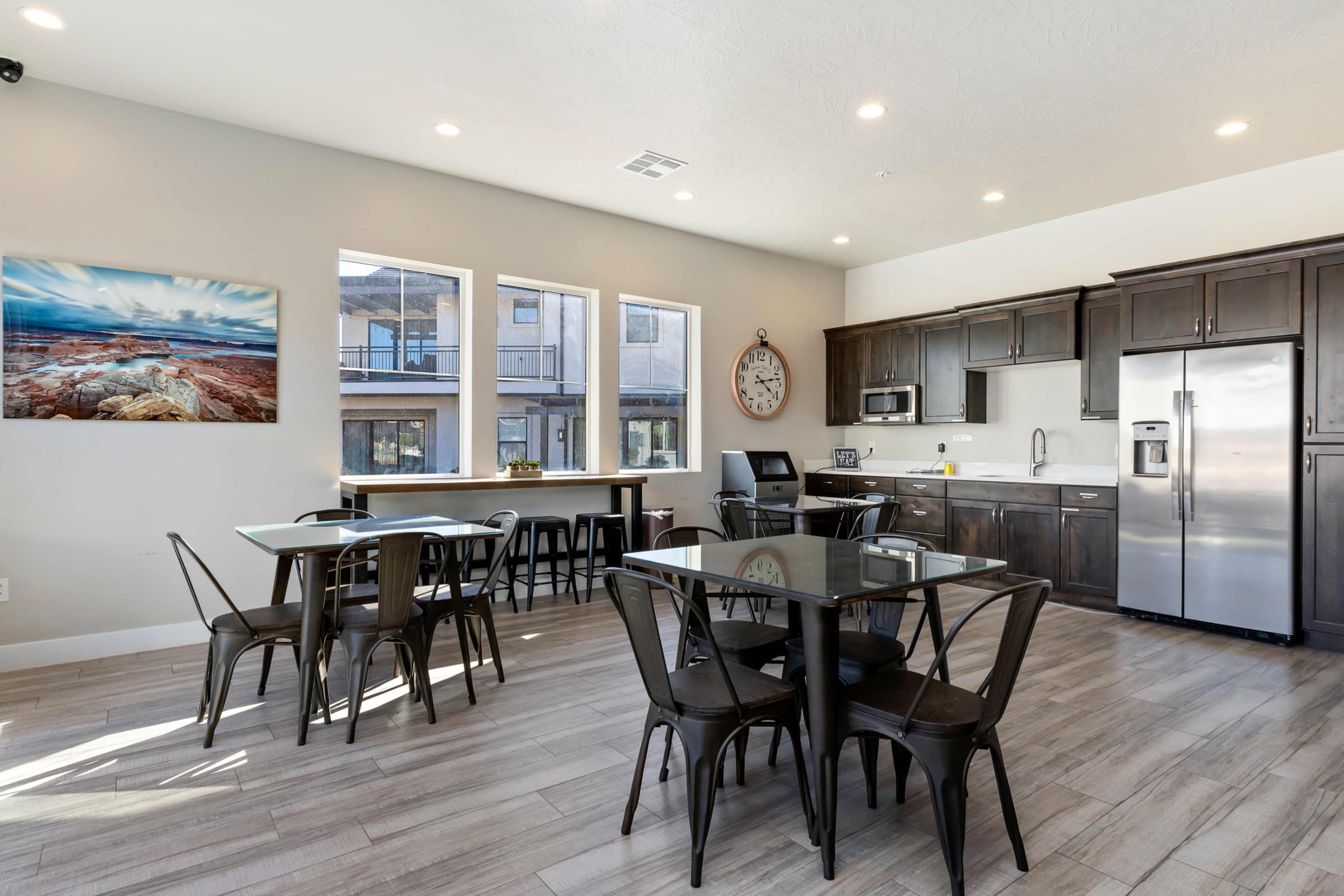Open-concept gathering space featuring wood-finish flooring, a full kitchen with dark cabinetry and stainless steel appliances, and a wall of three large windows