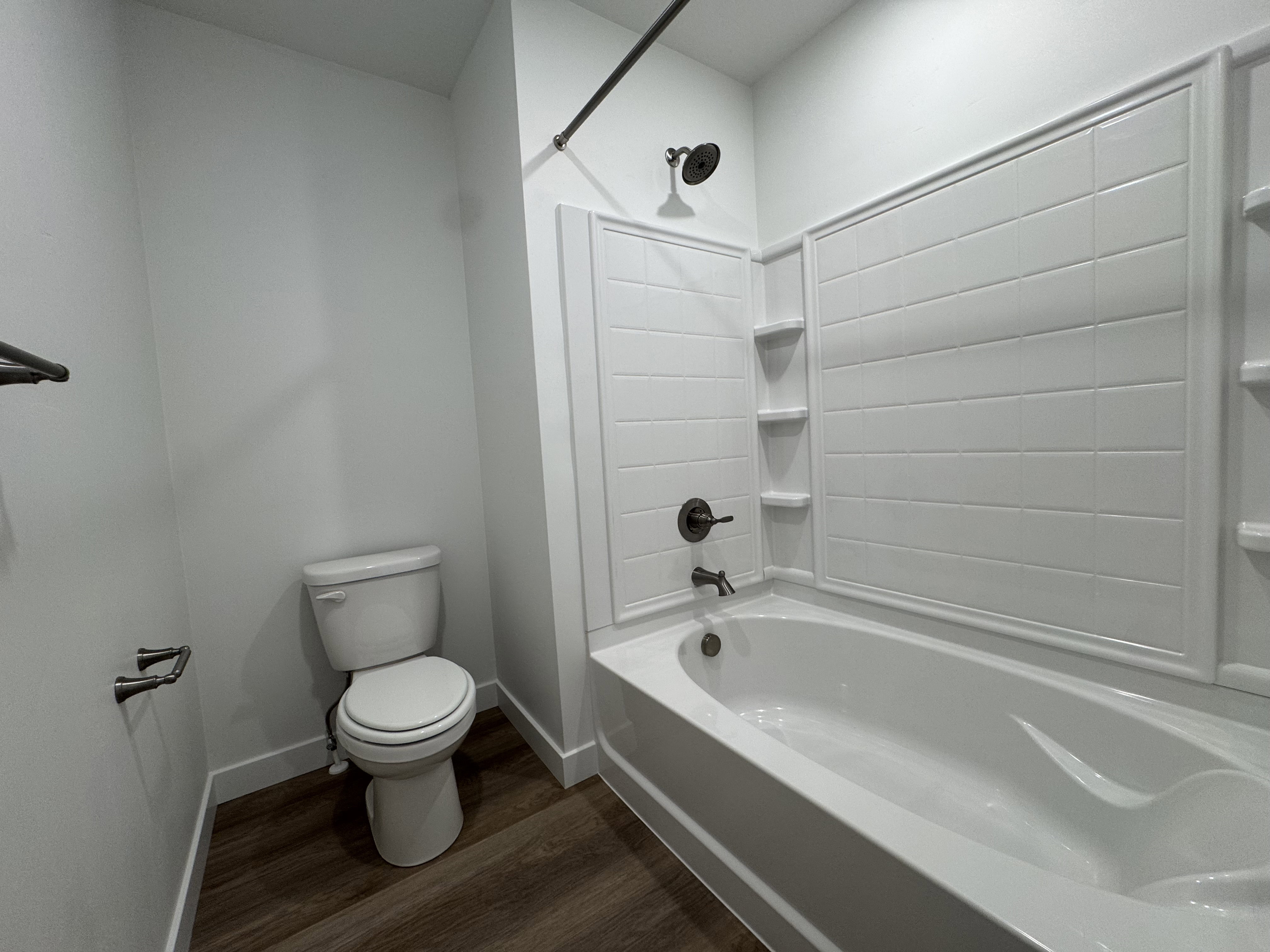 Bathroom featuring a white bathtub, white tiled shower surround with built-in shelving, dark bronze fixtures, and wood-finish flooring