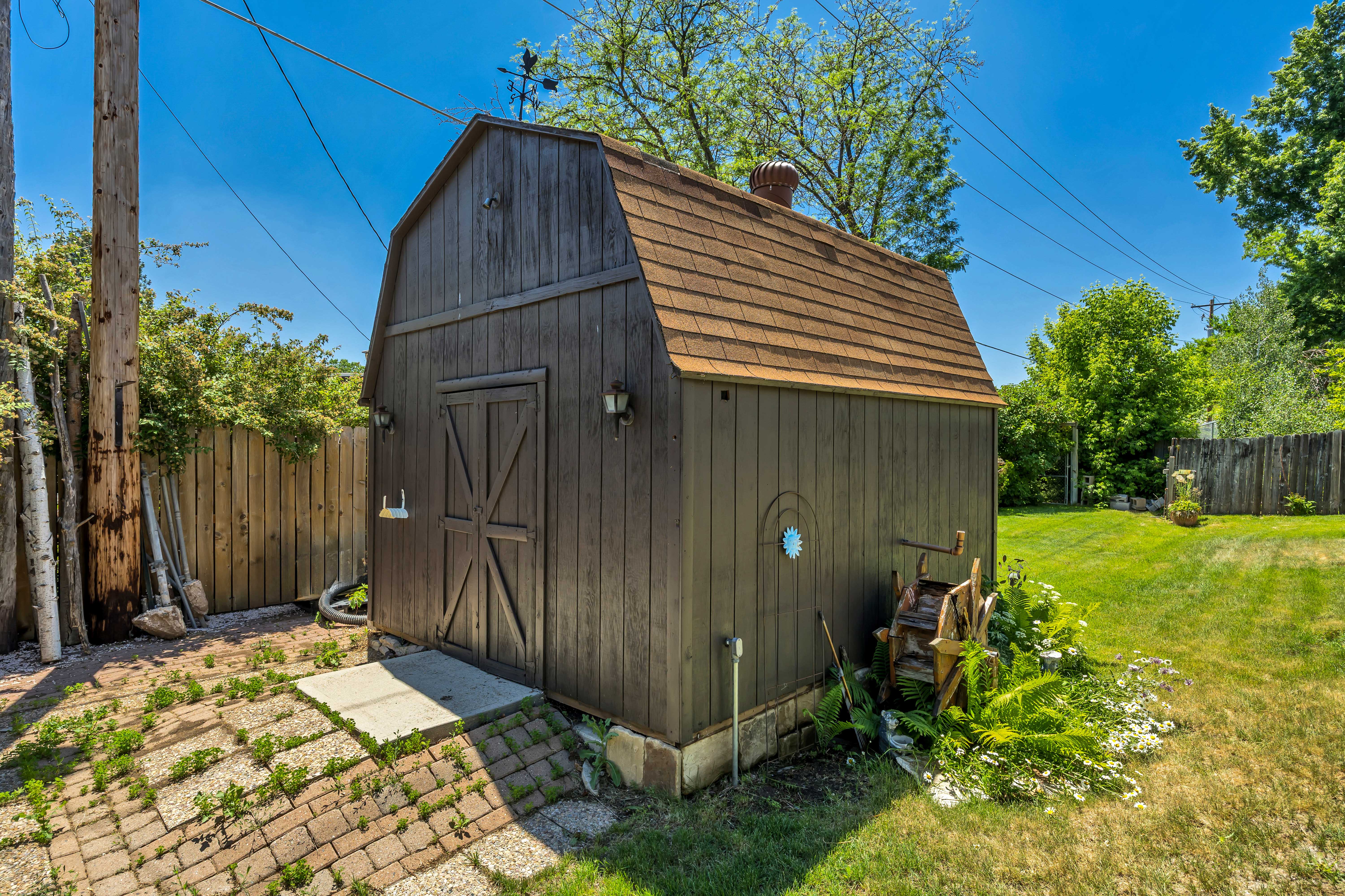 Freestanding barn-style shed featuring dark vertical siding, a shingled gambrel roof, and double barn doors