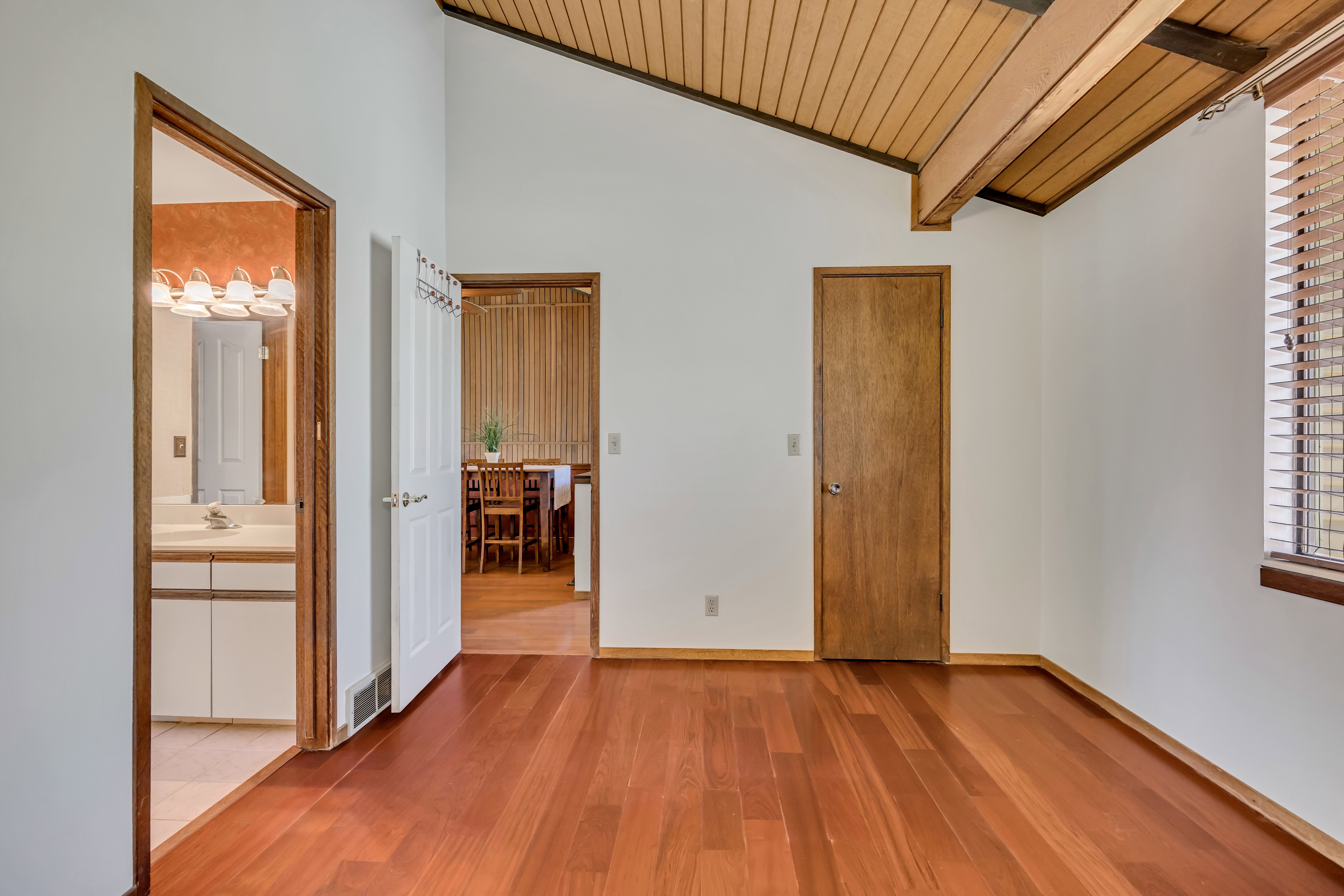 Interior space featuring wood-finish flooring, exposed wood beam ceiling, a window with horizontal blinds, and access to a bathroom with a vanity and an additional room with wood paneling