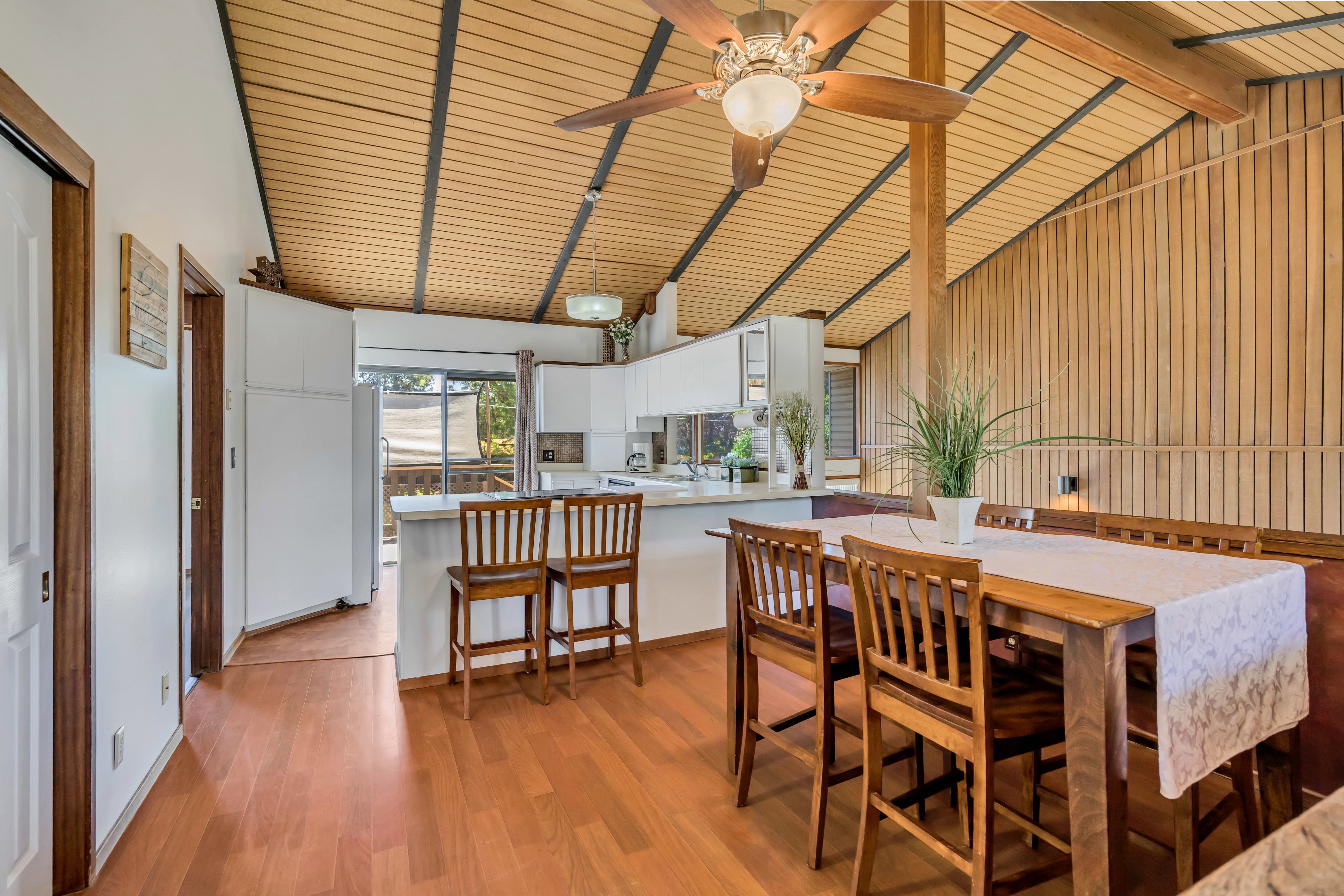 Vaulted wood-paneled ceiling with exposed beams, wood-finish flooring, kitchen with white cabinetry and solid surface countertops, ceiling fan, and sliding glass door leading to an exterior area