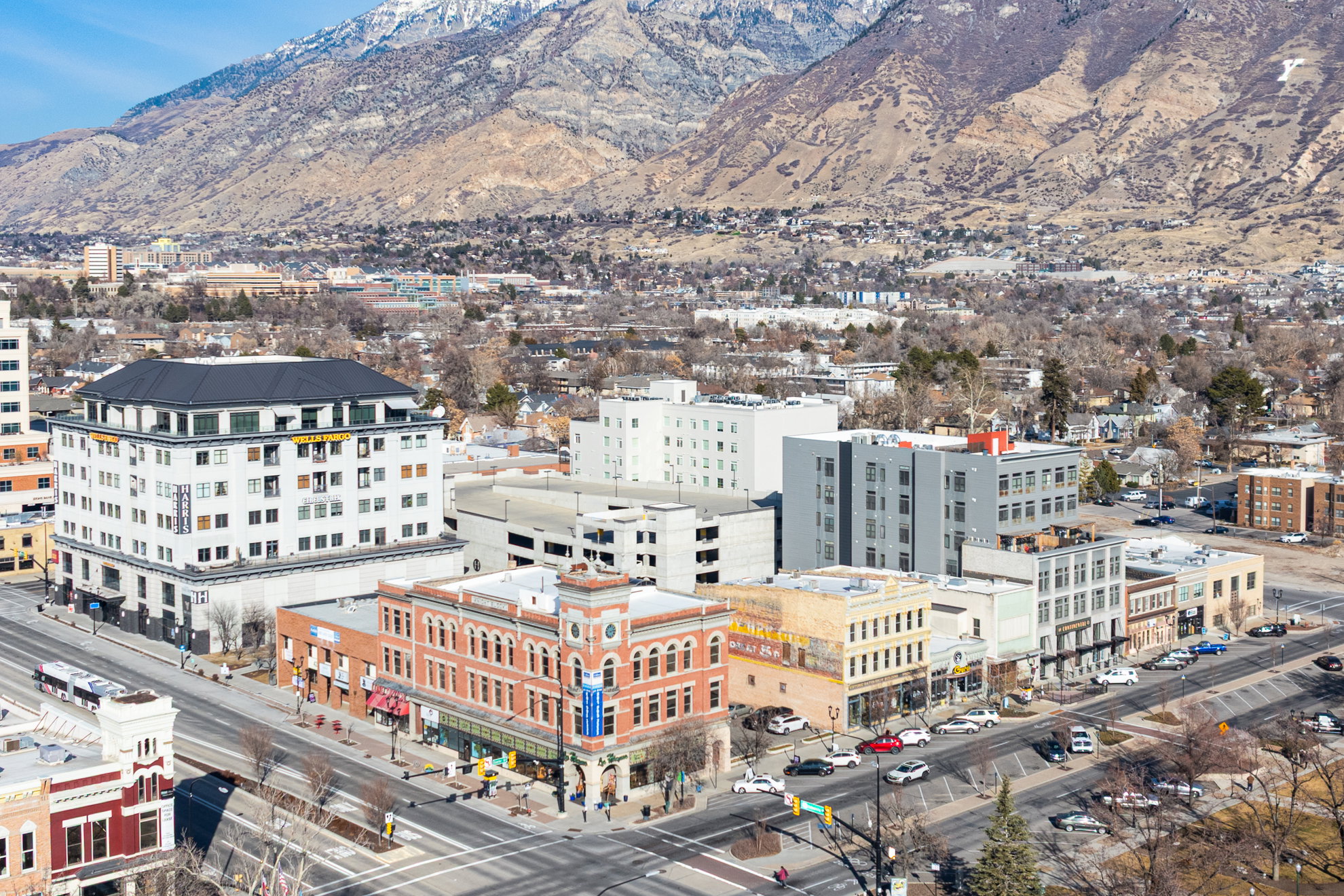 Urban landscape featuring diverse architectural styles, a prominent multi-story building with a dark roof, and a backdrop of mountains