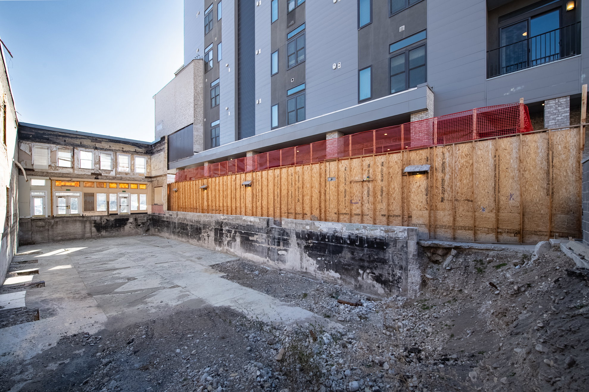 Concrete-paved ground area featuring surrounding concrete walls, a partial structure with multiple window openings, and a background building with dark gray siding and balconies