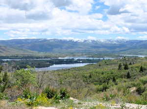 Expansive natural landscape with distant snow-capped mountains, a prominent lake, and rolling hills covered in diverse greenery