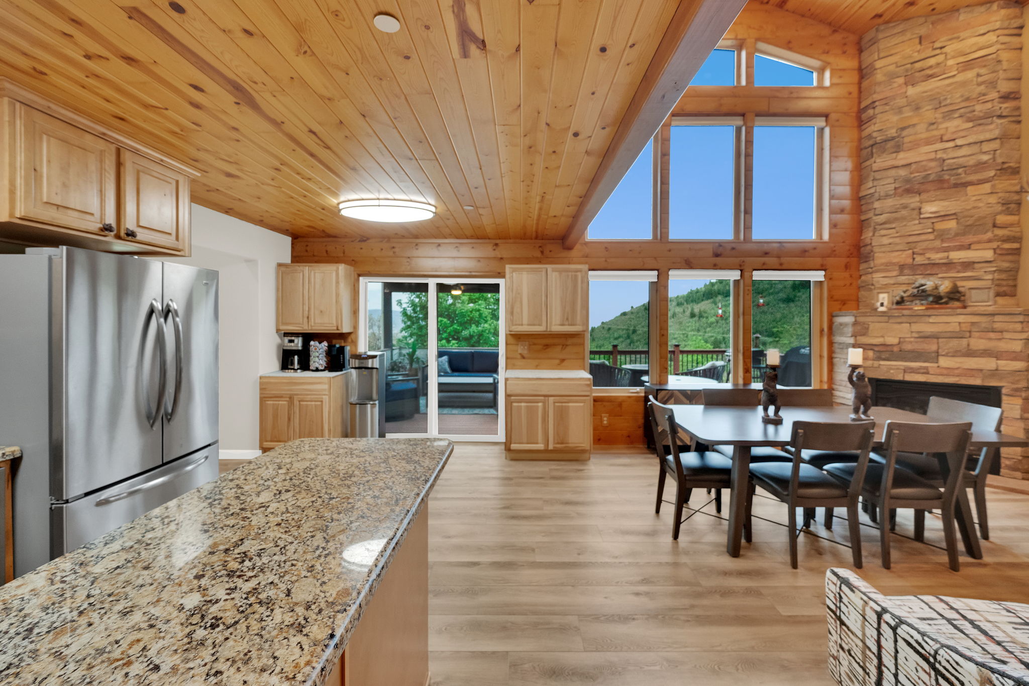 Open-concept living area featuring a vaulted wood-paneled ceiling, a stone-faced fireplace, a kitchen with granite countertops and stainless steel appliances, wood-finish flooring, and expansive windows showcasing a mountainous landscape