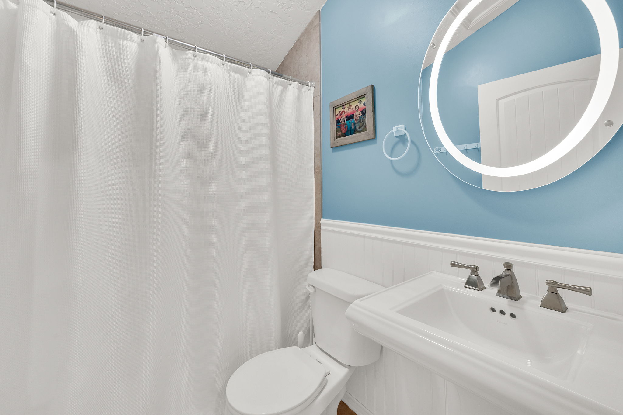 Bathroom featuring a pedestal sink with brushed nickel fixtures, an illuminated round mirror, white wainscoting, and a white toilet