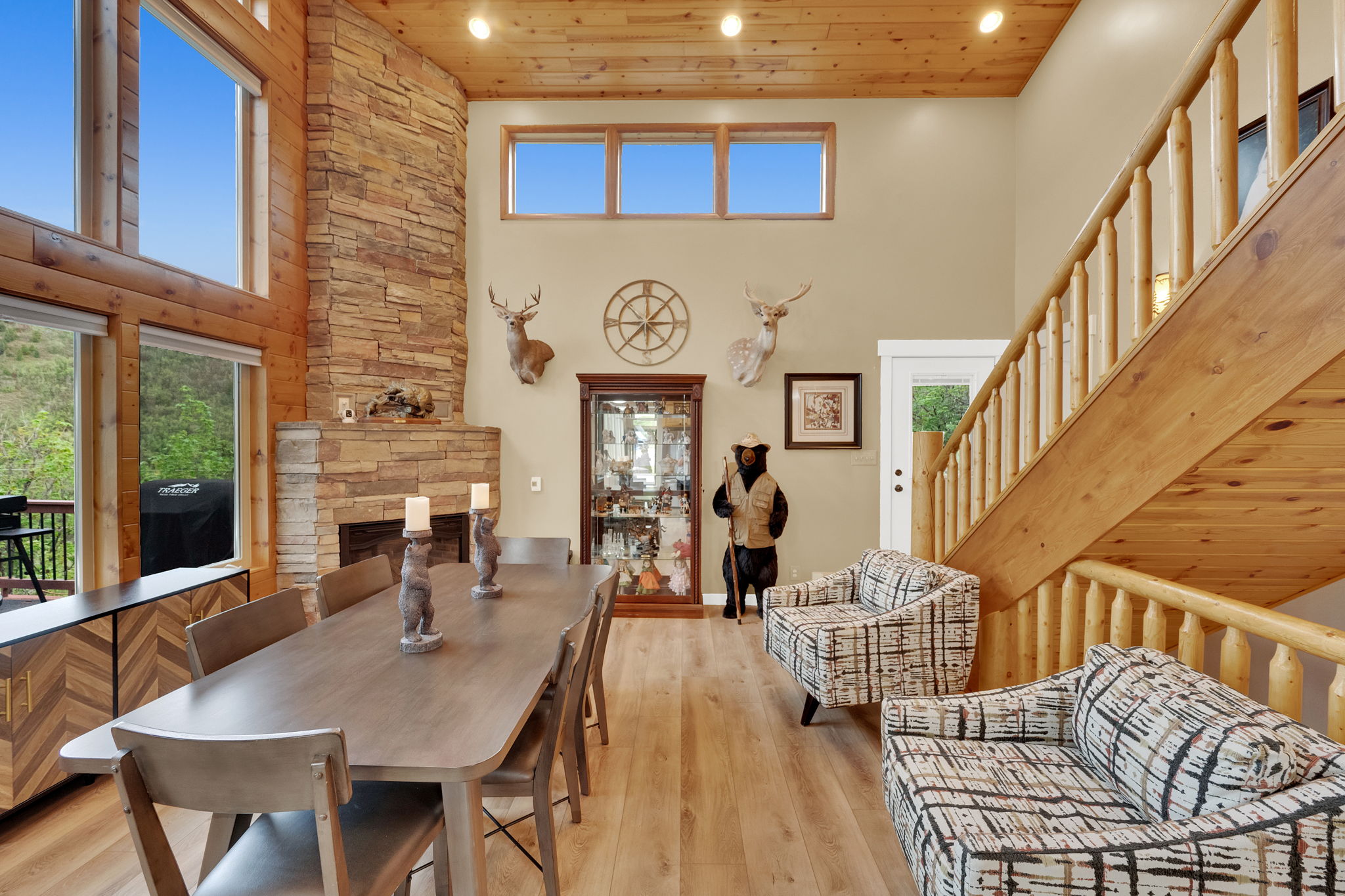 Spacious living area featuring a stone fireplace, wood-finish flooring, high ceilings, large windows, and an exposed wood staircase with log balusters