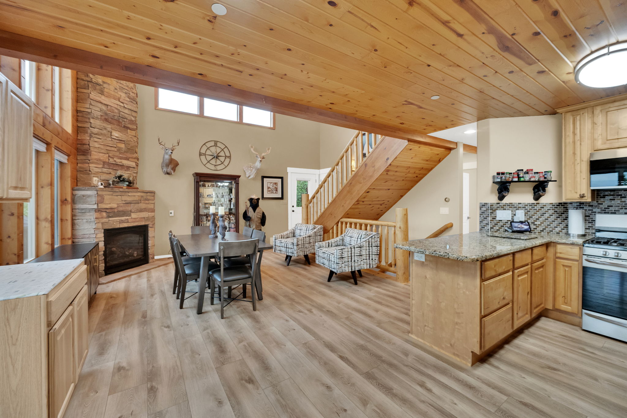 Open-concept living space featuring wood-finish flooring, a stone-faced fireplace, a natural wood paneled ceiling, a staircase with wood railings, and a kitchen island with a light-colored countertop