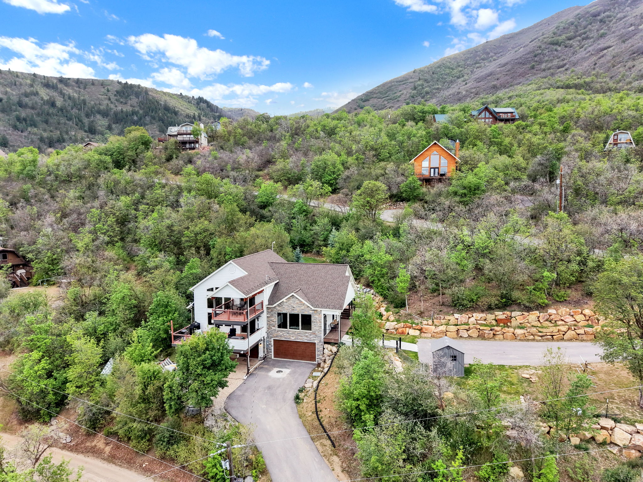 Custom mountain home featuring a stone and siding exterior, multi-level decks, an attached garage, and a winding paved driveway