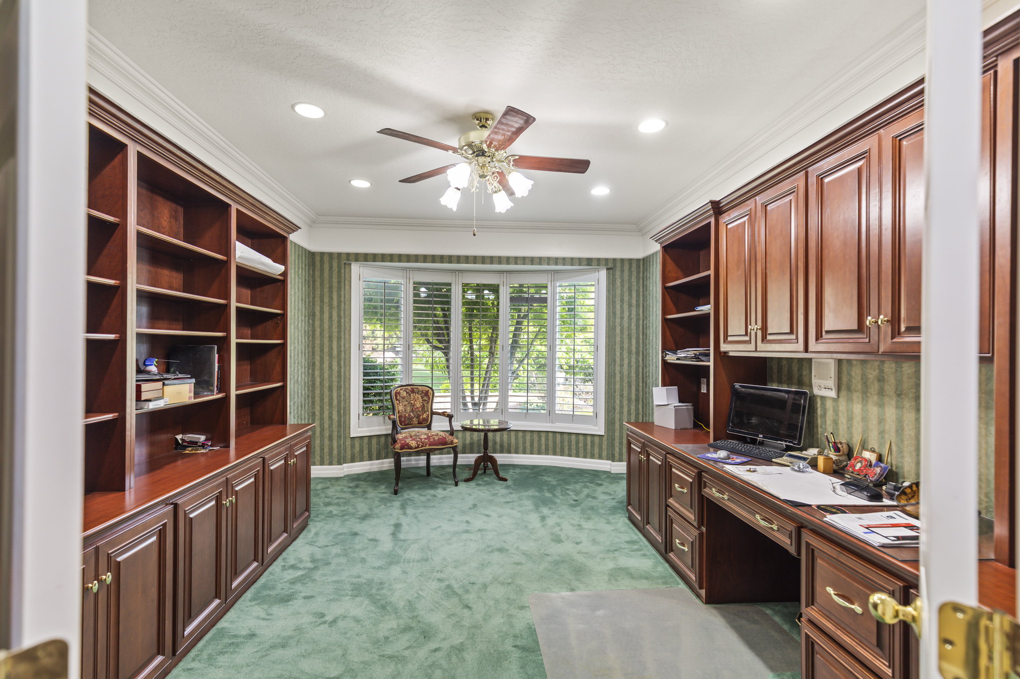 Office space featuring built in desk, wallpapered walls, ornamental molding, dark colored carpet, and a ceiling fan