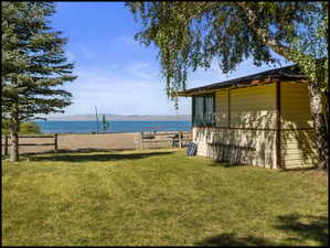 Waterfront property featuring a wooden fence bordering a sandy beach, a light-colored siding structure with a dark roof, a mature evergreen tree, and a deciduous tree