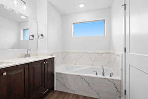 Bathroom featuring a built-in soaking tub with marble-look surround, a dual vanity with dark wood cabinetry and light countertops, modern light fixtures, and wood-finish flooring
