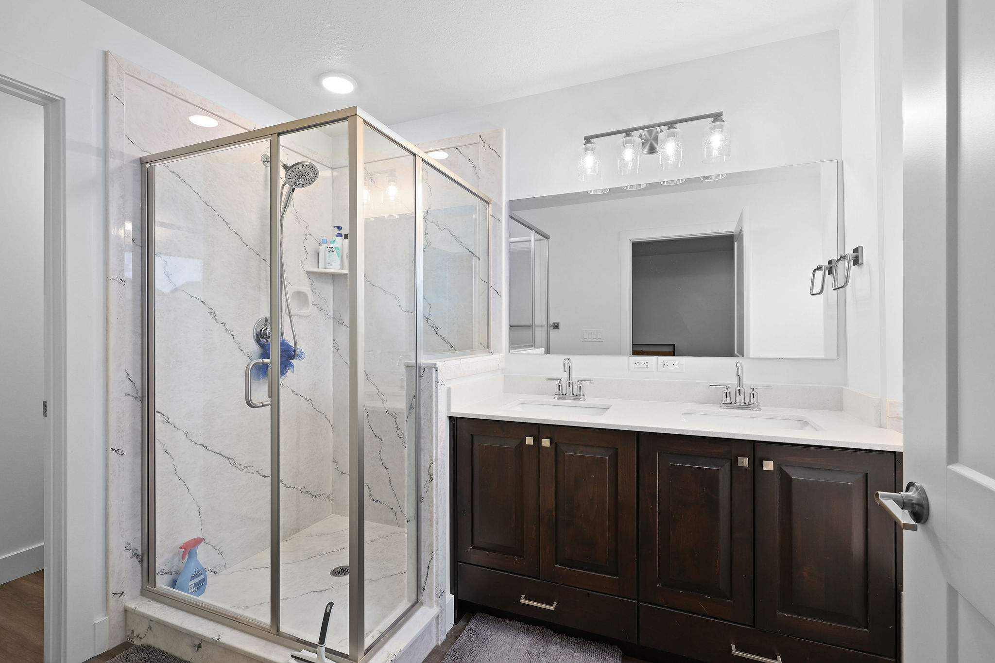 Spacious bathroom featuring a glass-enclosed shower with white veined tile surround, dual vanity with undermount sinks, dark wood-finish cabinetry, and a large mirror with a four-light fixture