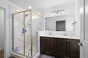 Spacious bathroom featuring a glass-enclosed shower with white veined tile surround, dual vanity with undermount sinks, dark wood-finish cabinetry, and a large mirror with a four-light fixture