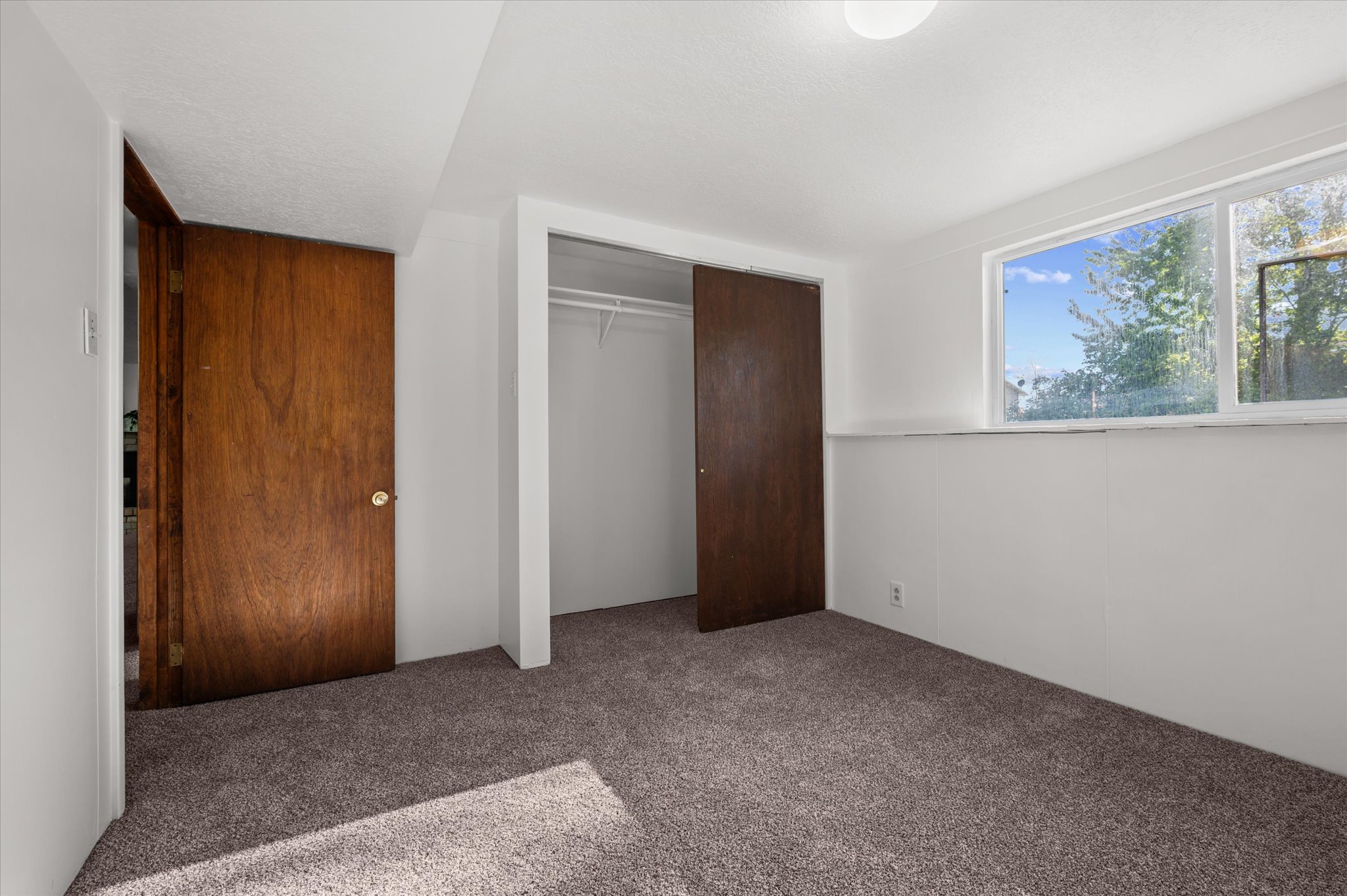 Carpeted room featuring a white ceiling and walls, a window with exterior views, a built-in closet with sliding wood doors, and an additional wood door