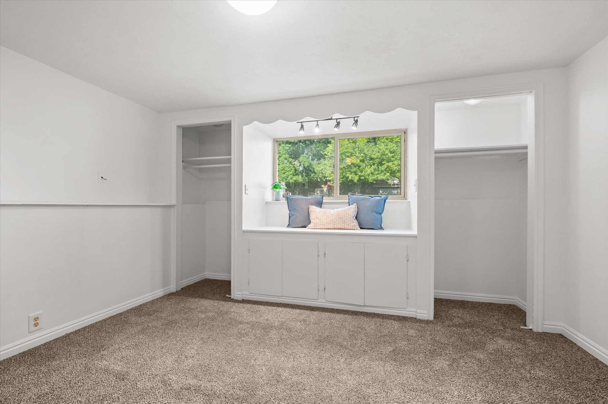 Built-in window seat with under-storage cabinetry, flanked by two closet alcoves, track lighting fixture, and neutral carpeting