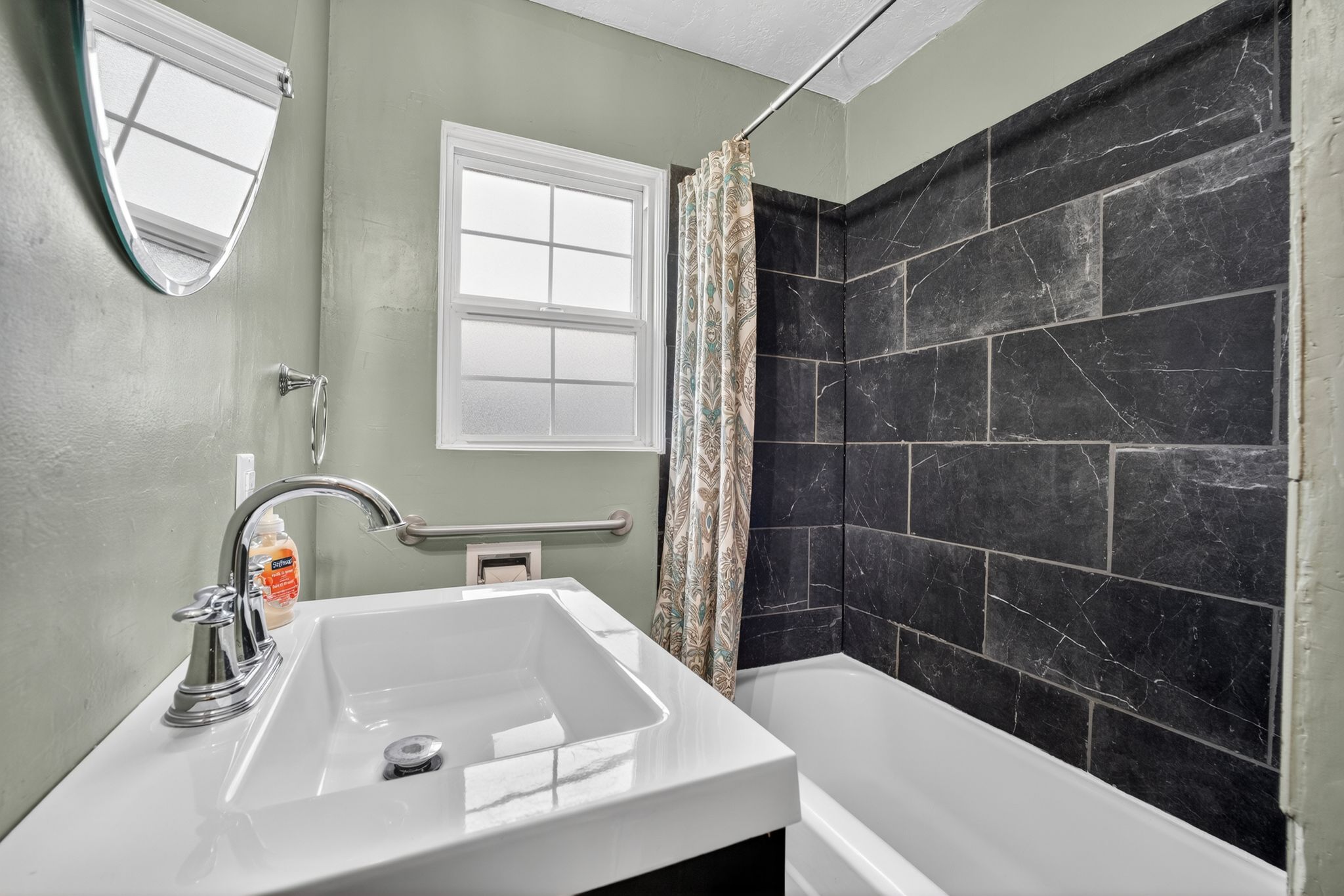 Full bathroom featuring a single white vanity with chrome faucet, a semi-oval wall-mounted mirror, a window with frosted glass, and a tiled shower-tub combination with dark, textured rectangular tiles