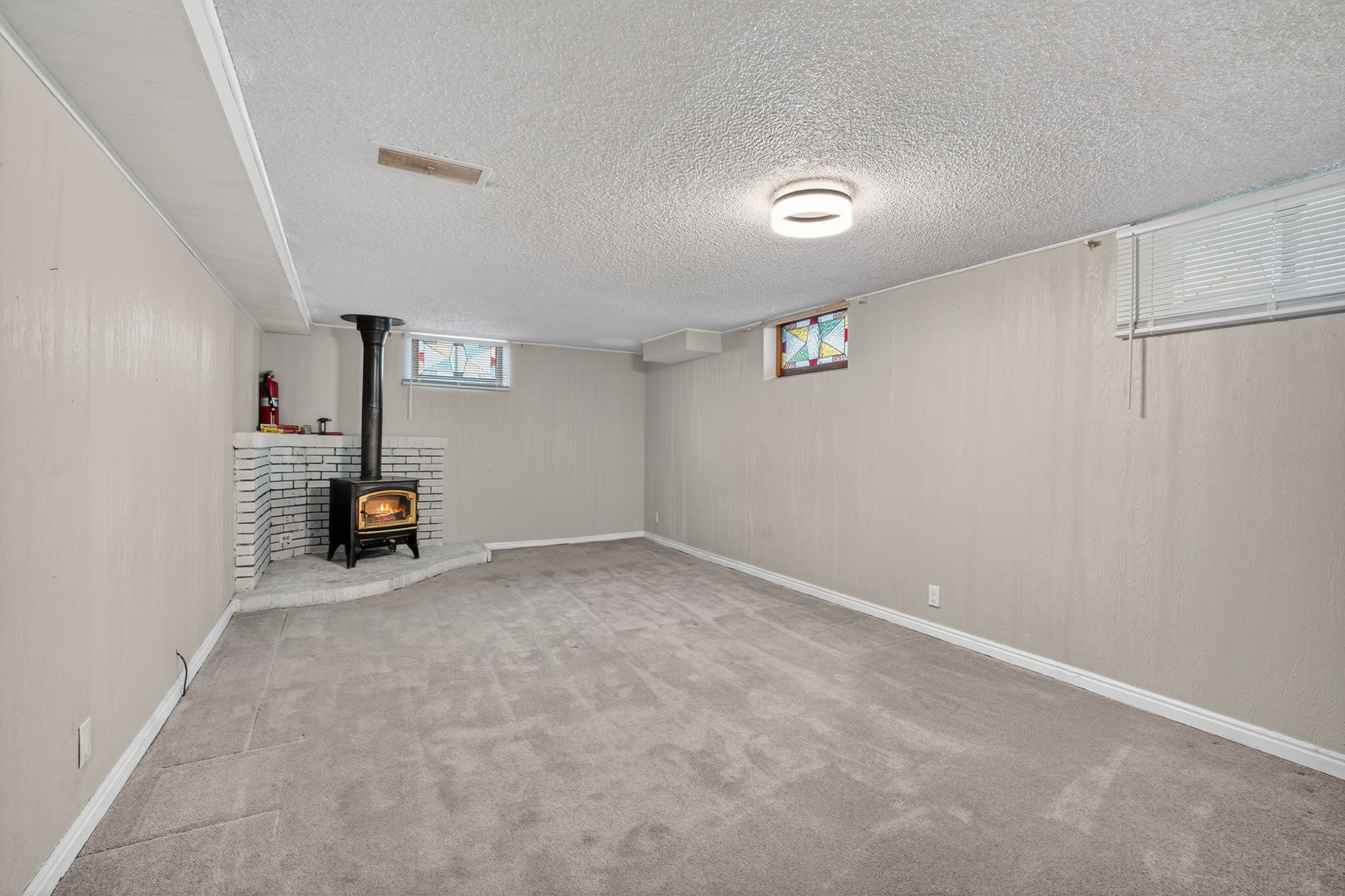 Spacious room featuring a corner wood-burning stove with a white brick surround and hearth, neutral carpeting, and a modern flush-mount ceiling light fixture