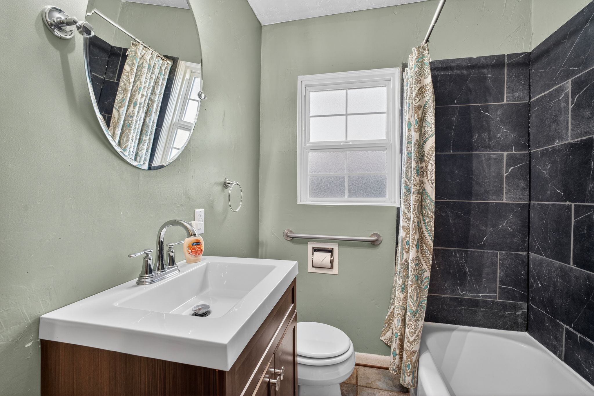 Bathroom featuring a dark wood-finish vanity with an integrated white sink, a modern oval mirror, a window with frosted glass panes, and a shower-over-tub with dark grey tiled walls