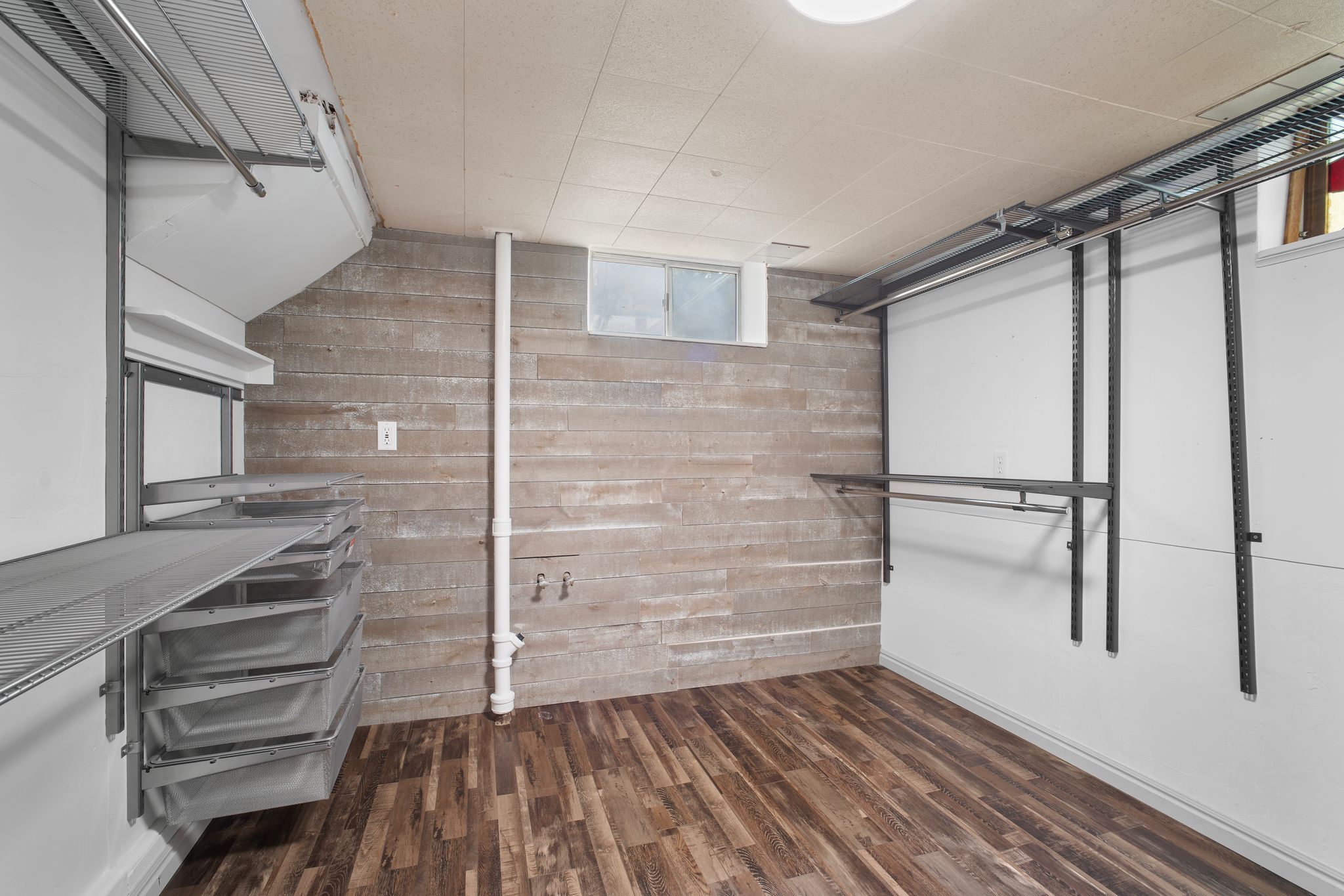 Versatile room featuring wood-finish flooring, a wood-paneled accent wall, a utility sink rough-in, an egress window, and built-in metal shelving