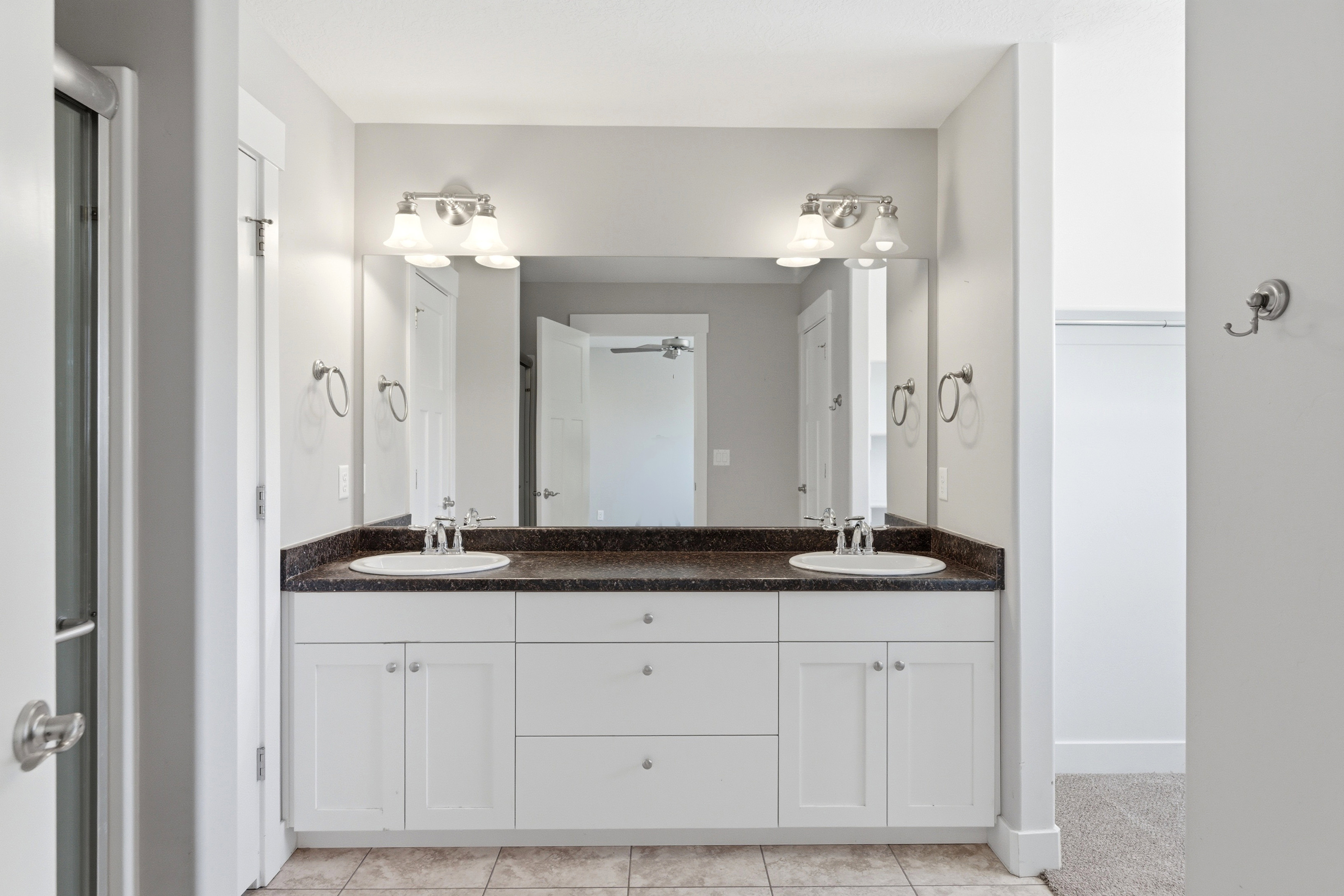 Bathroom vanity featuring dual under-mount sinks, white cabinetry, a dark speckled countertop, two wall-mounted vanity lights, and a large mirror