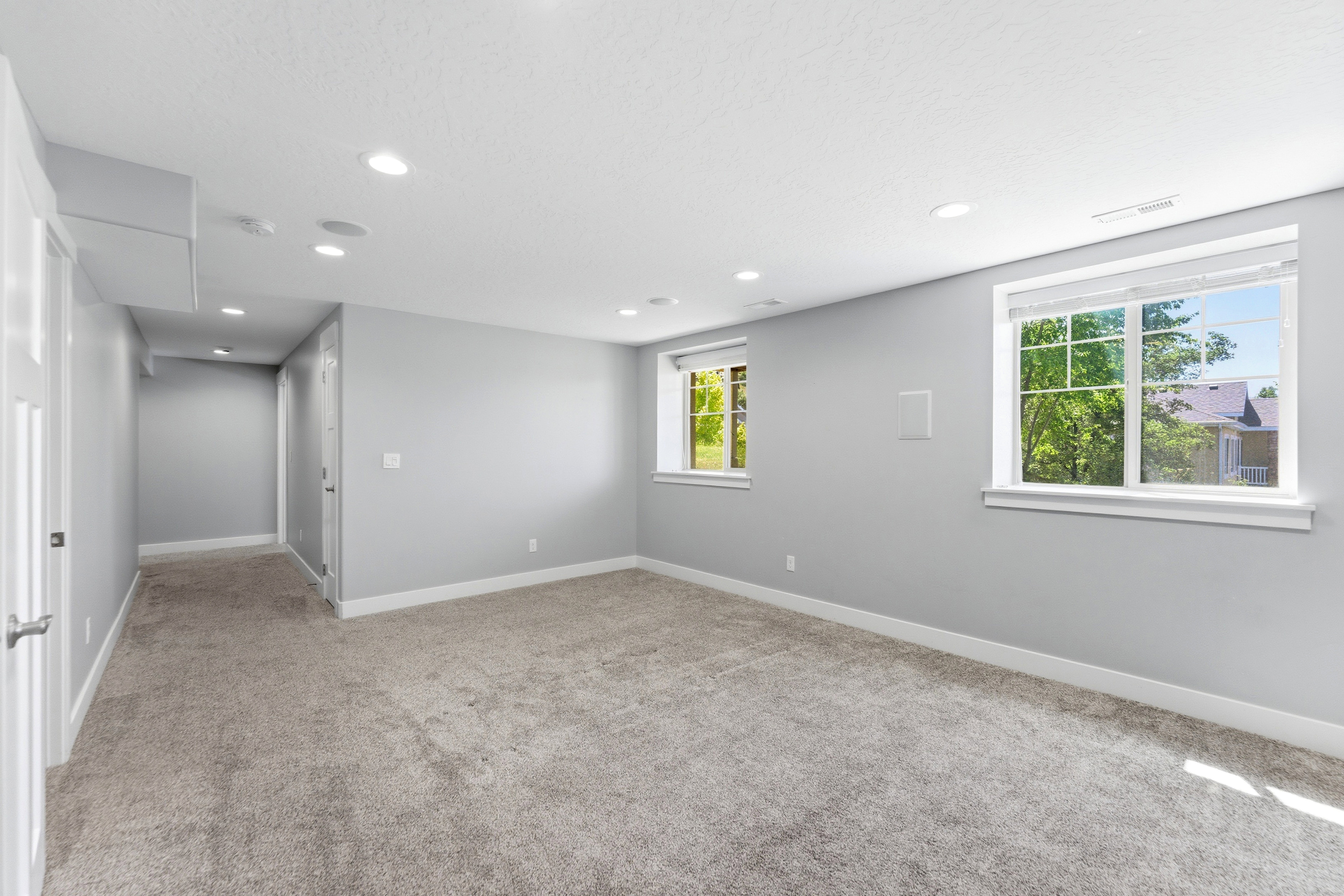 Spacious room featuring light gray walls, plush carpeting, multiple recessed lights, and two windows with white trim