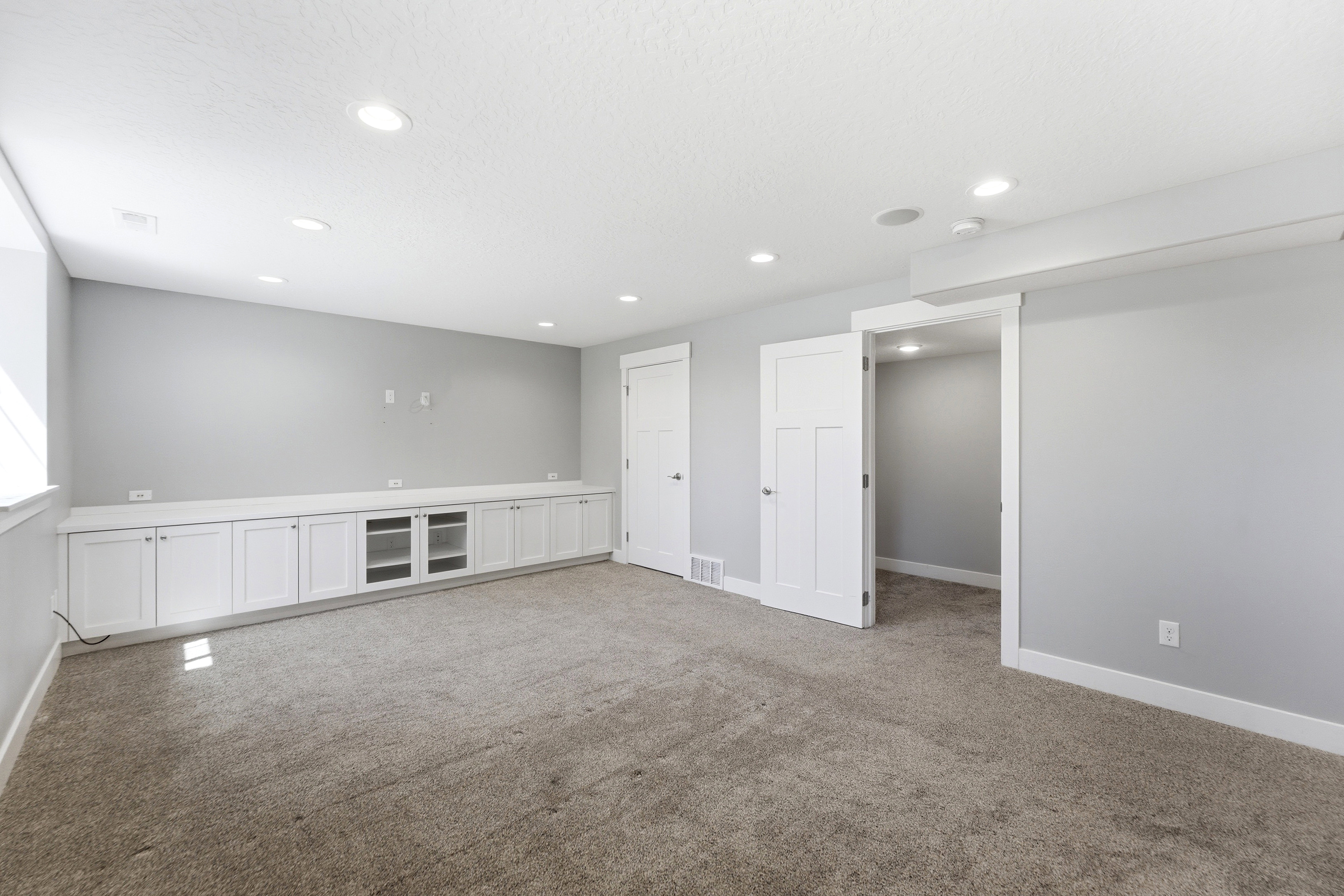 Spacious room featuring light gray walls, recessed lighting, wall-to-wall carpeting, and an extensive built-in media console with white cabinetry and display shelving