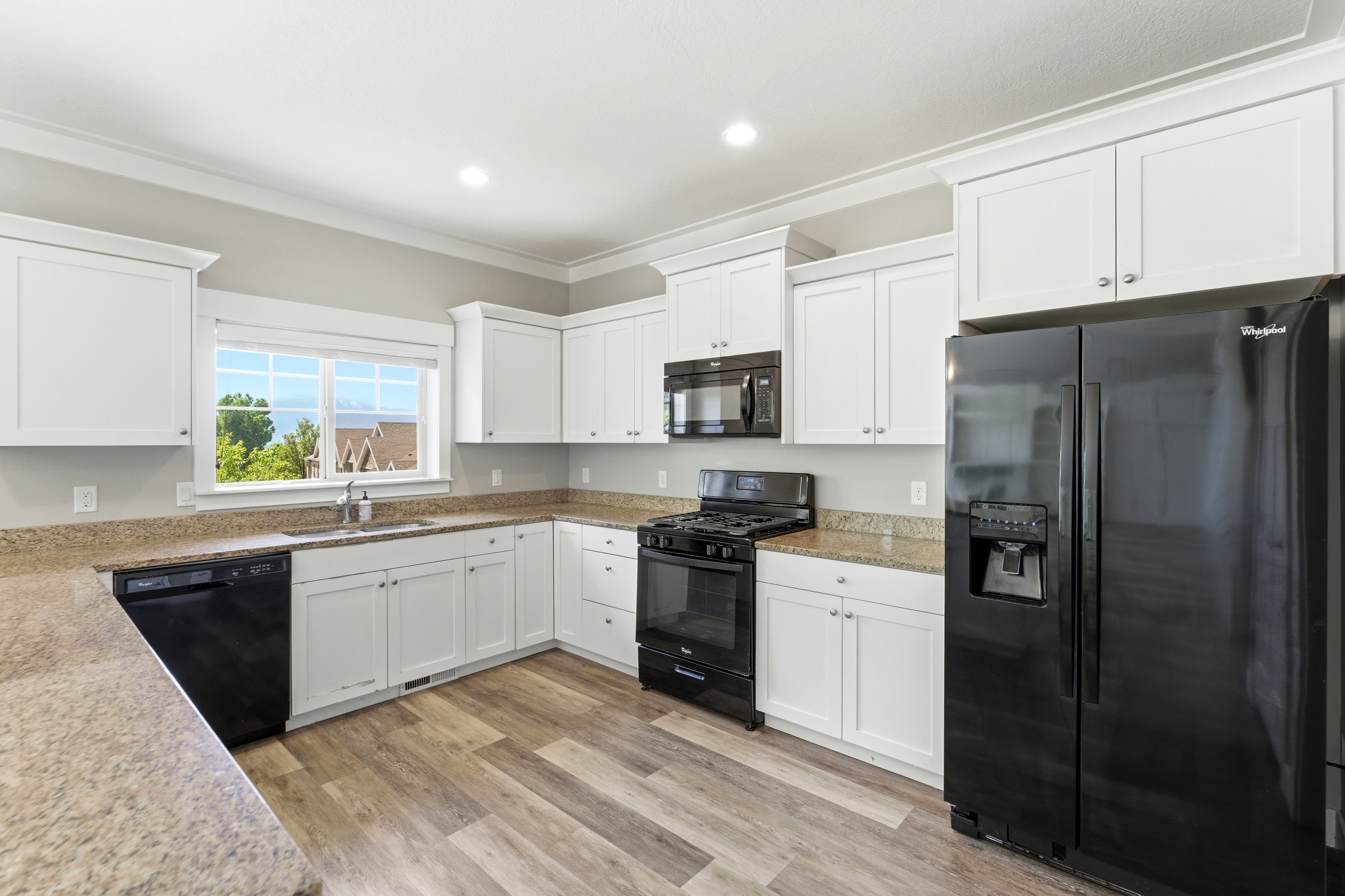 Modern kitchen featuring white shaker cabinetry, granite-finish countertops, black appliance suite, wood-finish flooring, and a large window above the sink