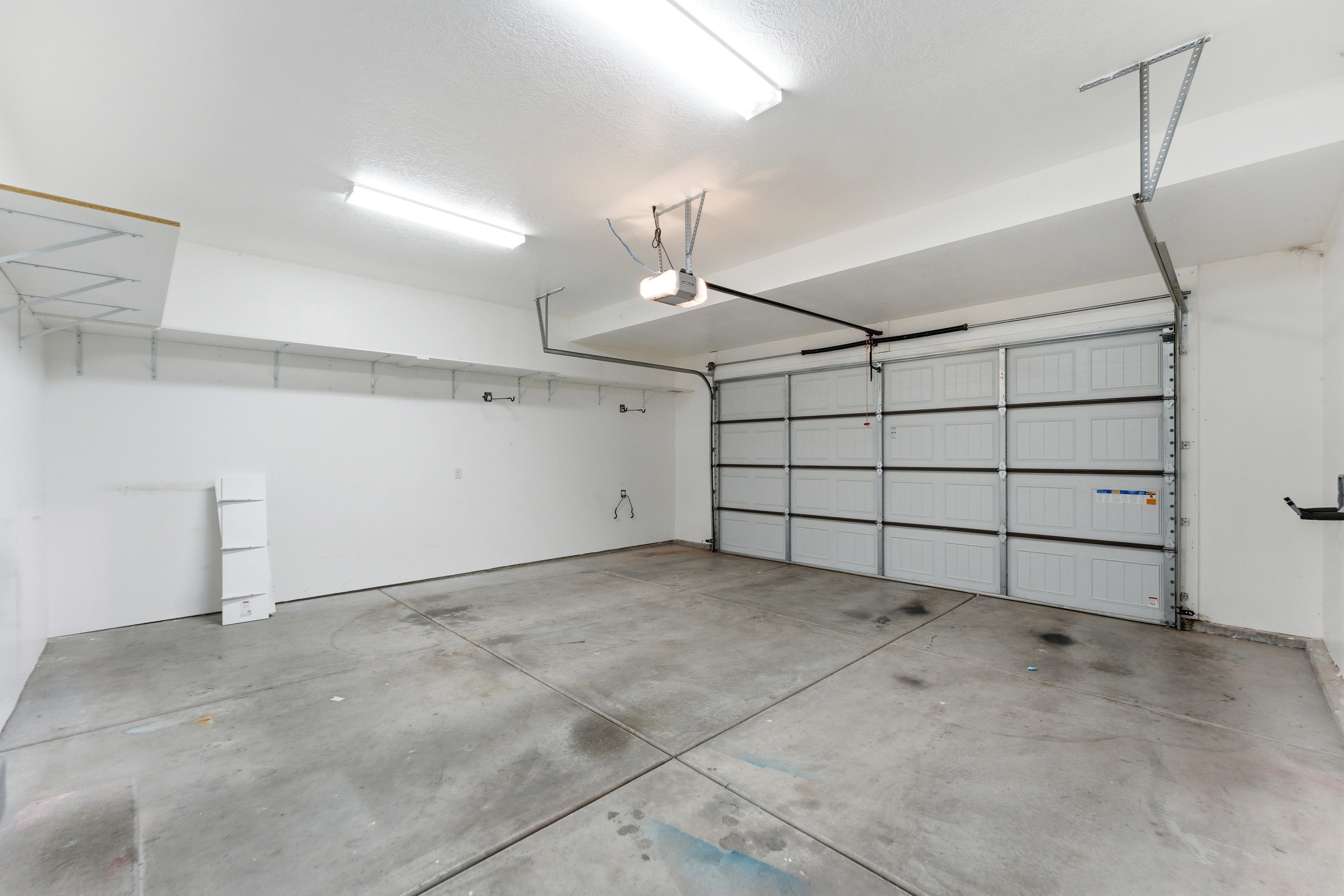 Spacious two-car garage with a panel garage door, concrete flooring, overhead fluorescent lighting, and built-in wall shelving