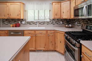 Kitchen featuring wood cabinetry, light-toned countertops, a mosaic tile backsplash, stainless steel appliances, and tile flooring